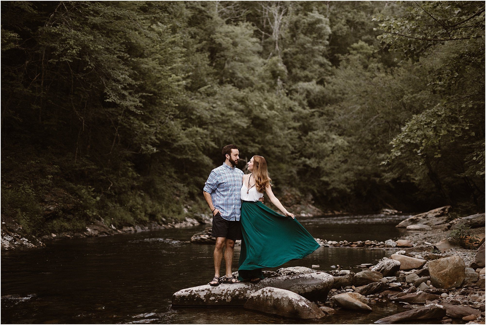 girl holding green skirt while looking at man standing on rocks in the middle of river