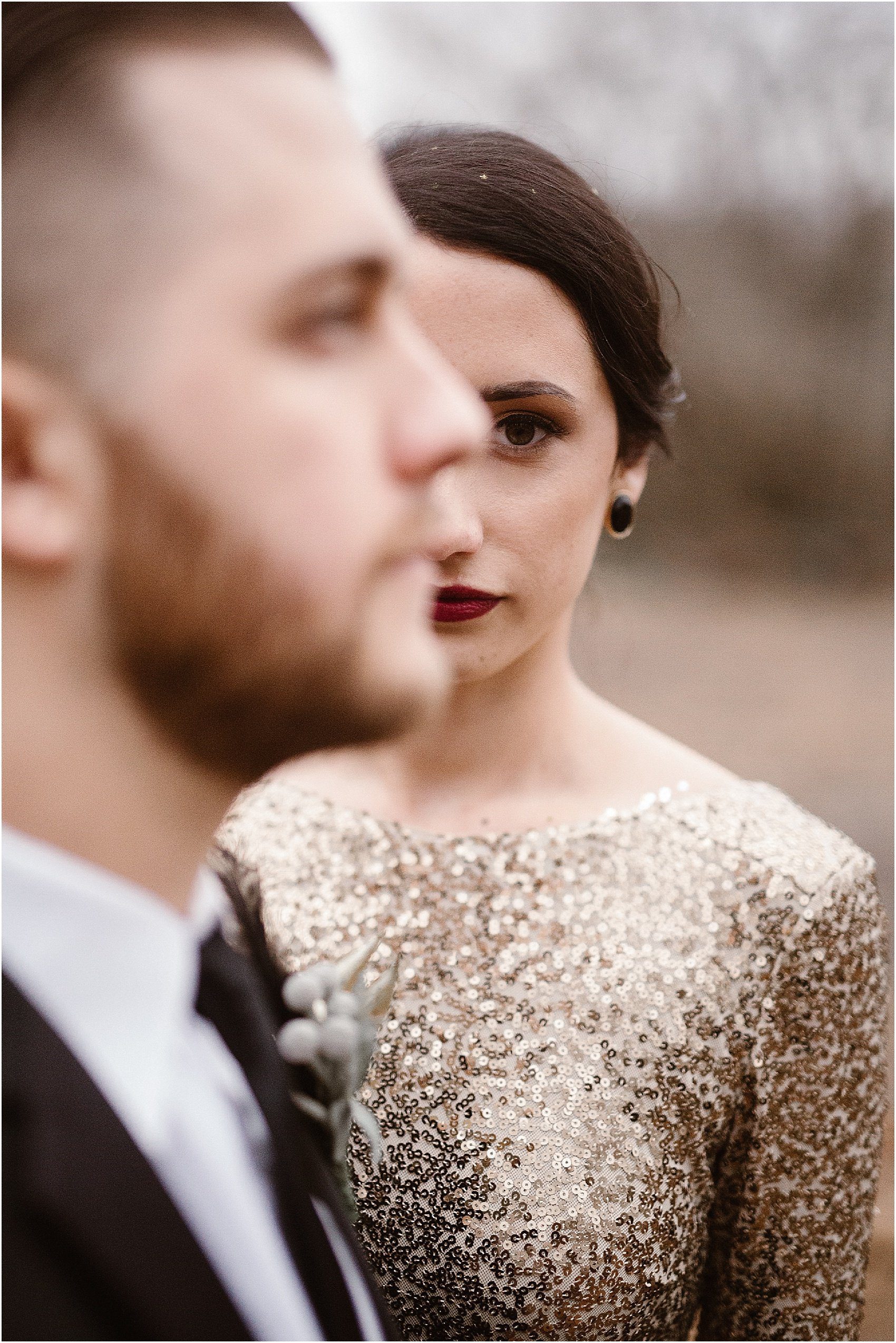 woman stands behind man wearing black earrings and gold wedding dress