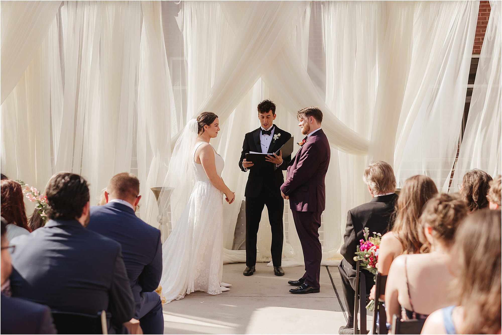 A bride and groom stand facing each other at an outdoor wedding ceremony, with an officiant between them and guests on both sides. Sheer white drapes create a soft, elegant backdrop—moments like these show why you need a professional wedding photographer.