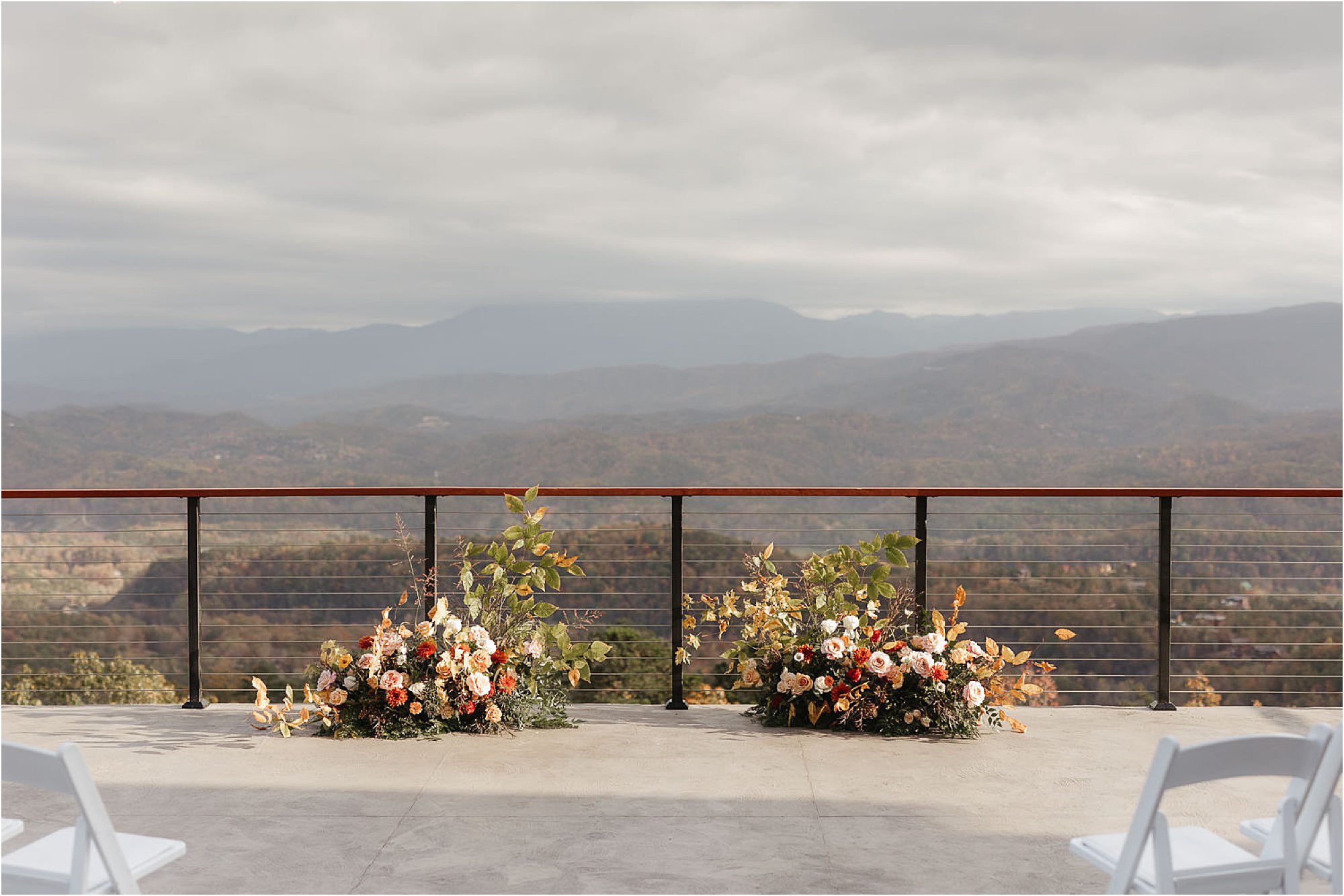 Floral arrangements with peach and cream flowers sit on a patio overlooking distant mountains, creating a scene that shows why you need a professional wedding photographer to capture every stunning detail. White chairs are visible in the foreground.