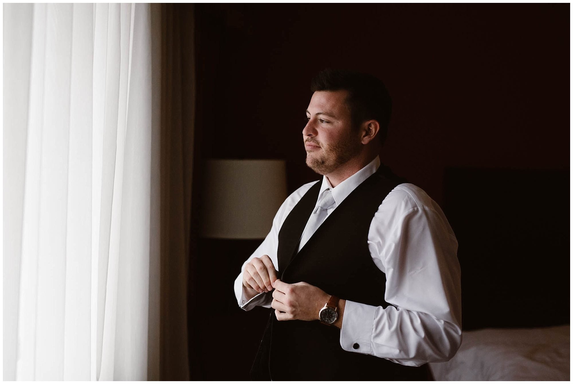 groom putting on vest in front of window at hotel