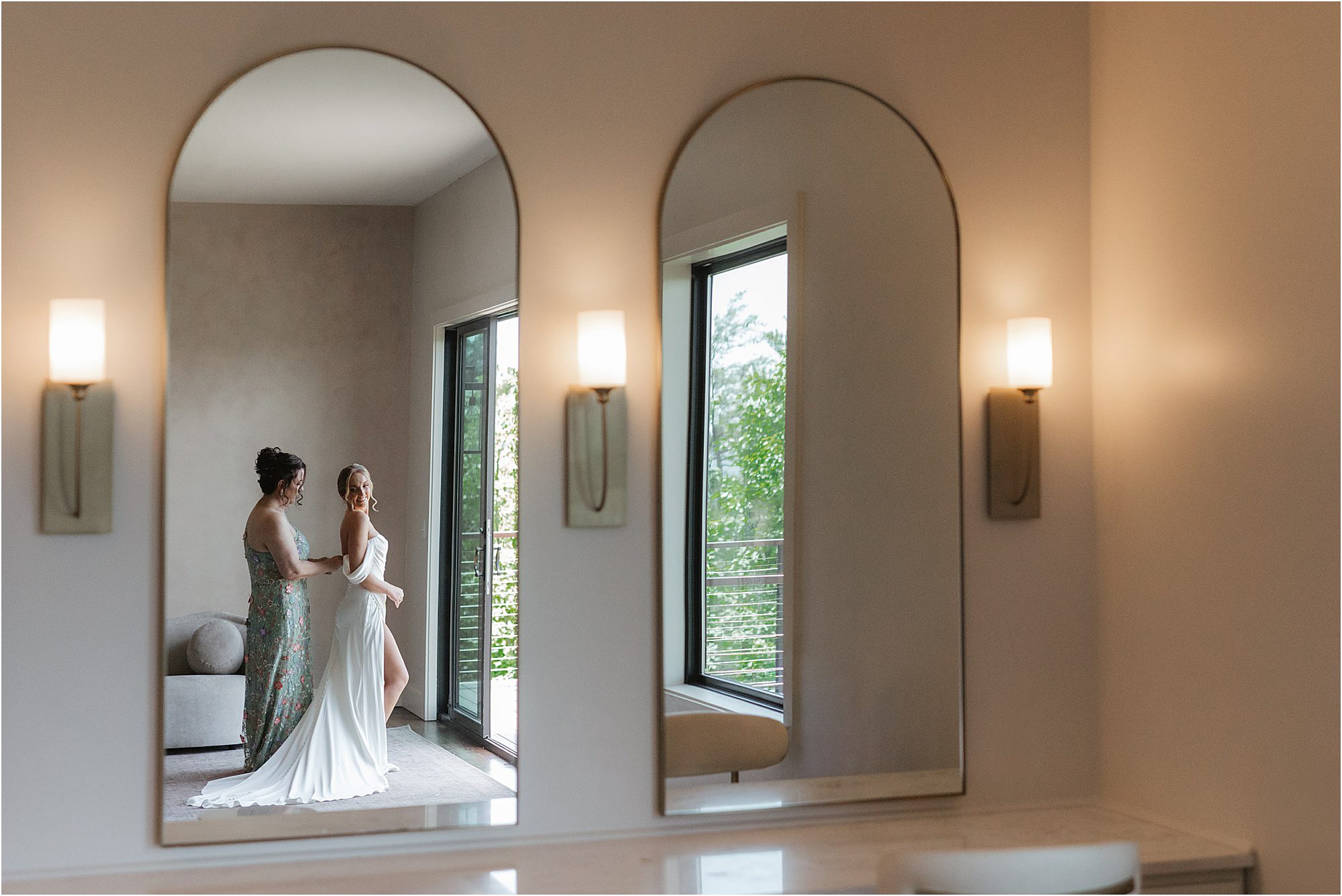 Two women, one in a white dress and one in a floral dress, stand together and smile, reflected in a large mirror in a softly lit modern room with wall sconces and large windows showing greenery outside—a perfect scene that captures some of the best tips for getting ready on your wedding day.