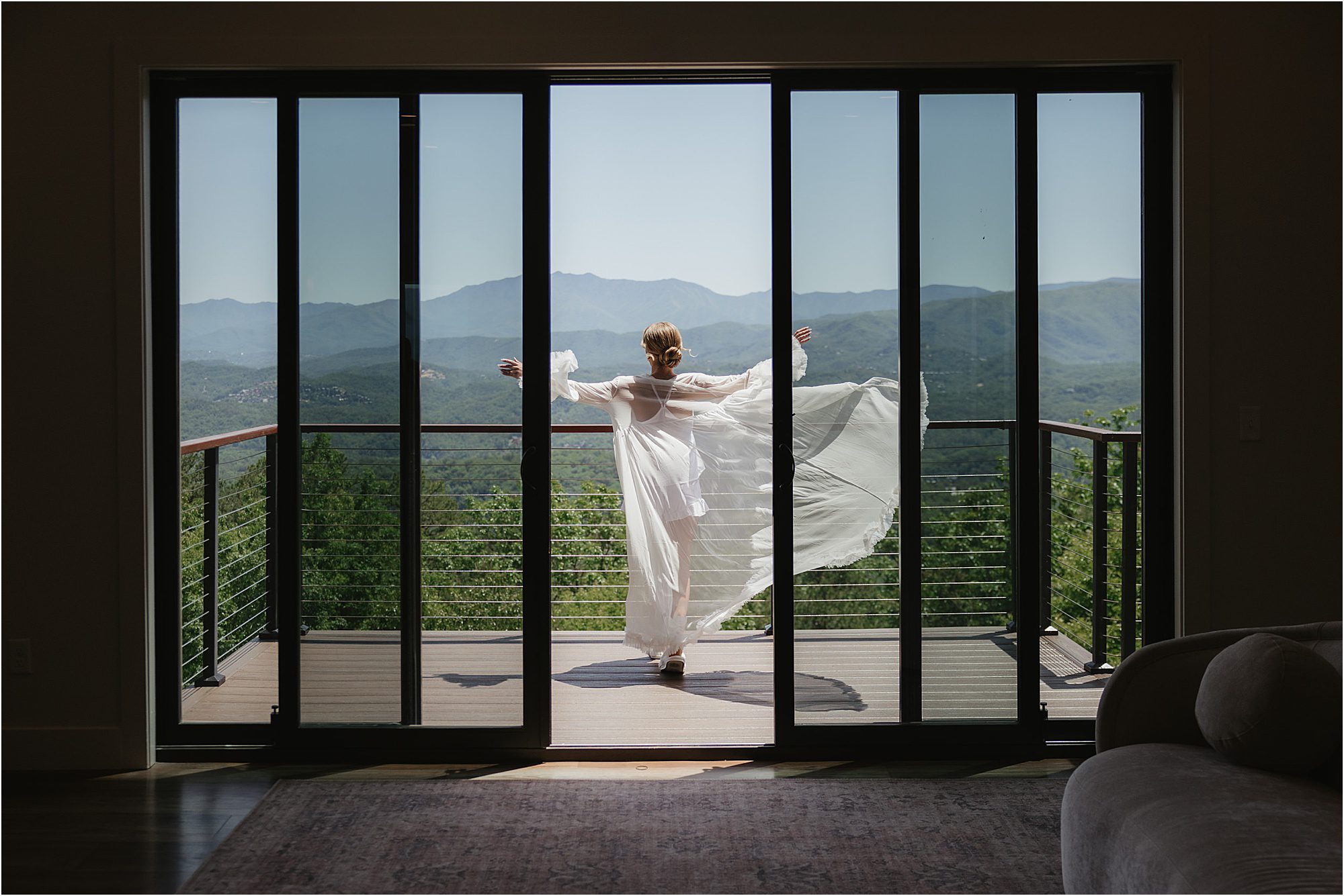 A person in a flowing white dress stands on a balcony with arms outstretched, facing a scenic mountain landscape, as seen through open glass doors—capturing one of those serene moments often cherished in tips for getting ready on your wedding day.
