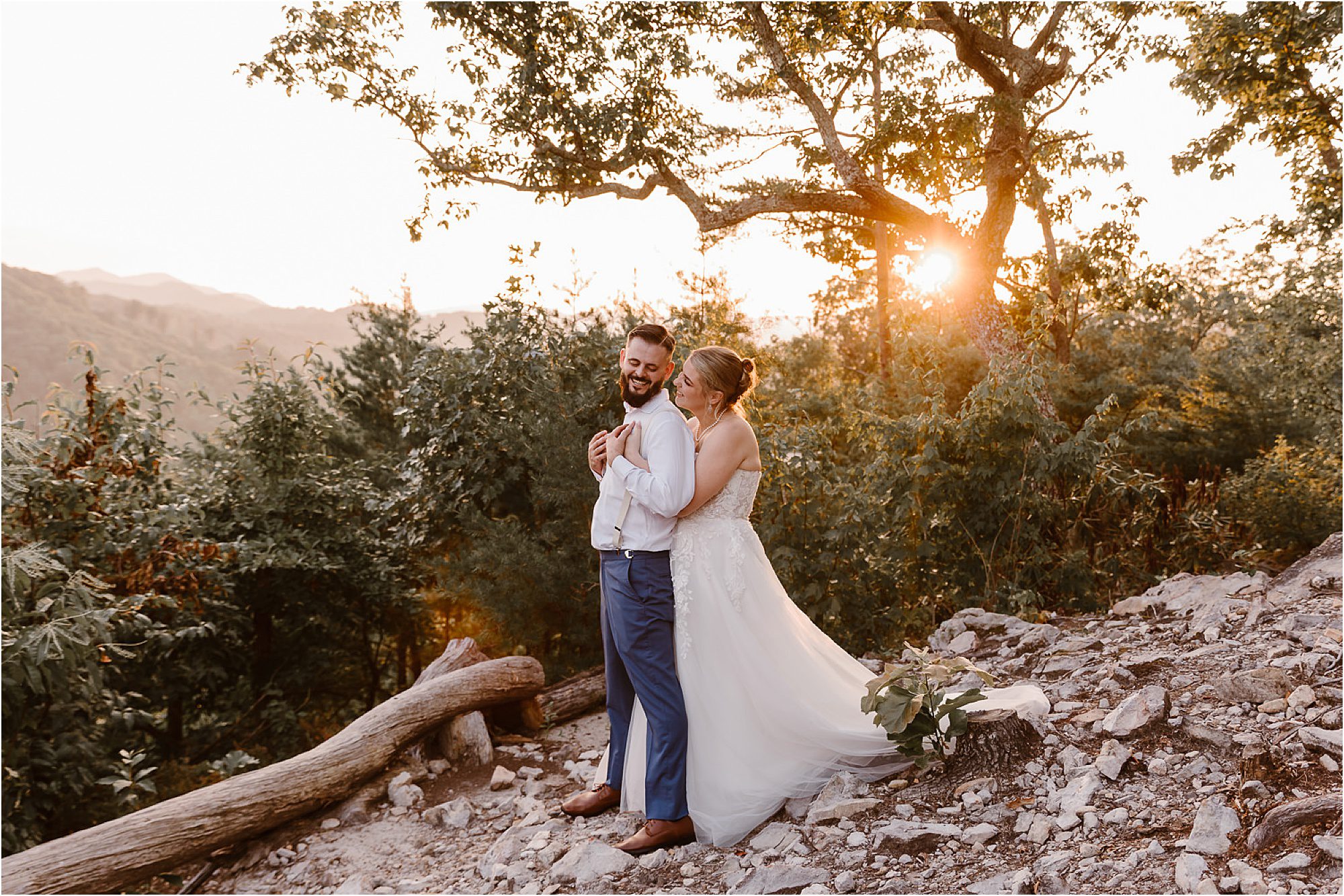 A bride and groom embrace on a rocky hillside at sunset, surrounded by trees and mountains. Sunlight streams through the trees behind them, casting a dreamy glow—perfect for golden hour photography.
