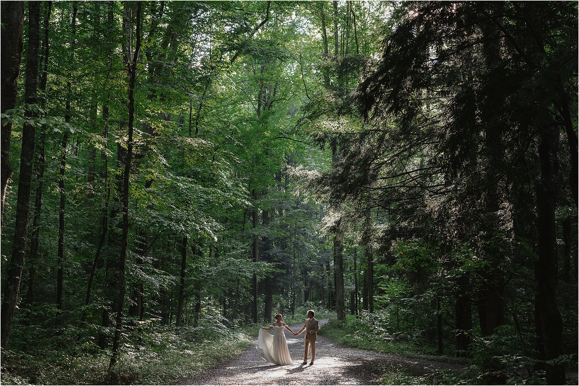A couple in wedding attire holds hands while walking on a sunlit, tree-lined gravel path through a dense forest in Great Smoky Mountain National Park. Lush green foliage surrounds them, and soft sunlight filters through the trees, creating a magical setting for their Elkmont Photos.