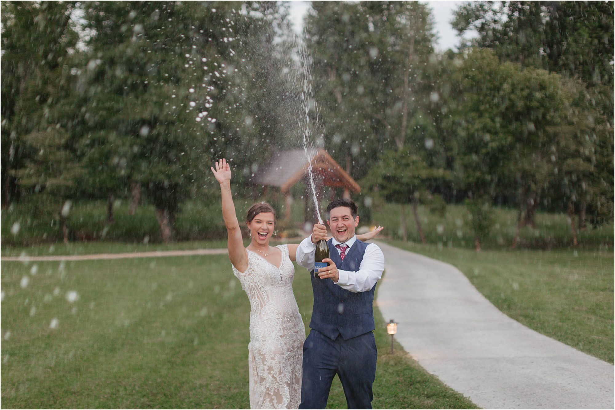 A joyful bride and groom celebrate outdoors, smiling and spraying champagne—highlighting the festive spirit of alcohol at weddings—with trees and a pathway in the background.