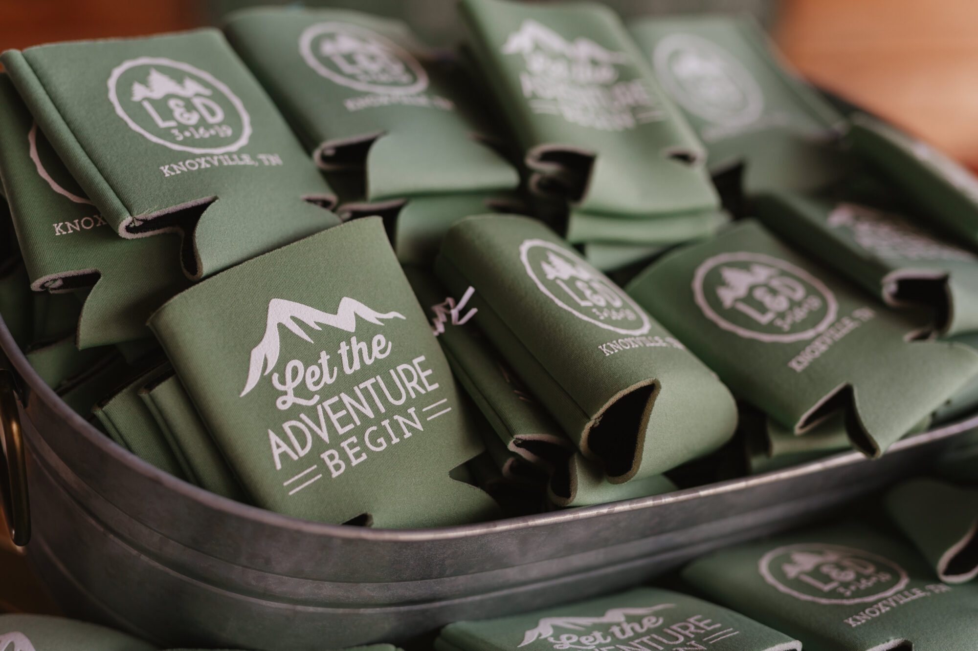 A metal container brimming with green can koozies adorned with white designs. The text says, Let the Adventure Begin, next to a mountain graphic, perfect for wedding receptions. The initials L&D and Knoxville, TN add a touch of personalization to these charming keepsakes.