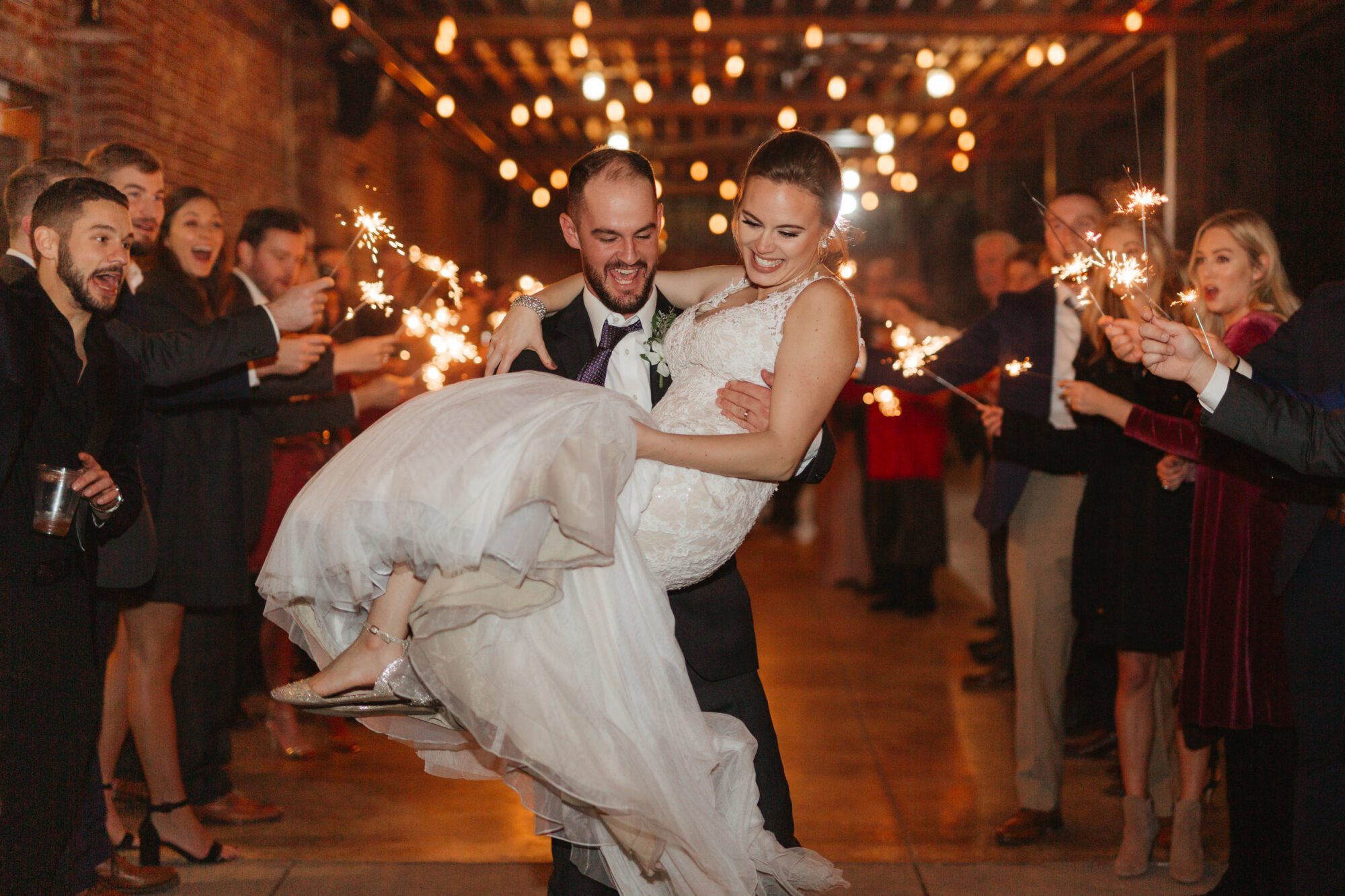 A bride in a white dress is joyfully carried by the groom in a dark suit, surrounded by a cheering crowd holding sparklers. Under string lights in a warmly lit space, they create unforgettable memories—truly one of the tips for a successful wedding reception.