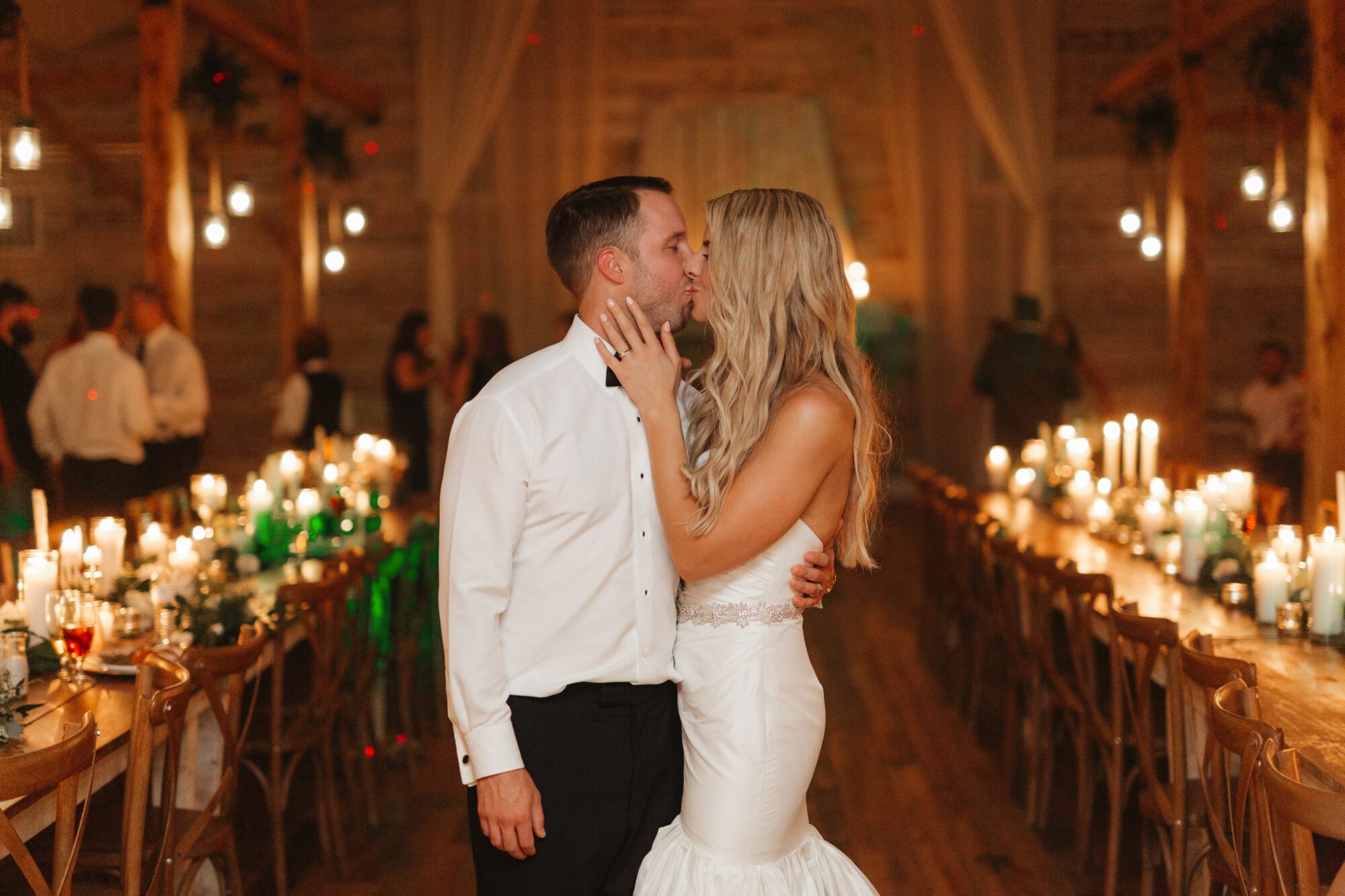 A bride and groom share a kiss in a warmly lit reception hall, capturing one of many tips for a successful wedding reception. Long tables with candles and floral arrangements line the wooden room. The bride wears a white gown, and the groom is in a white dress shirt. Guests are blurred in the background.