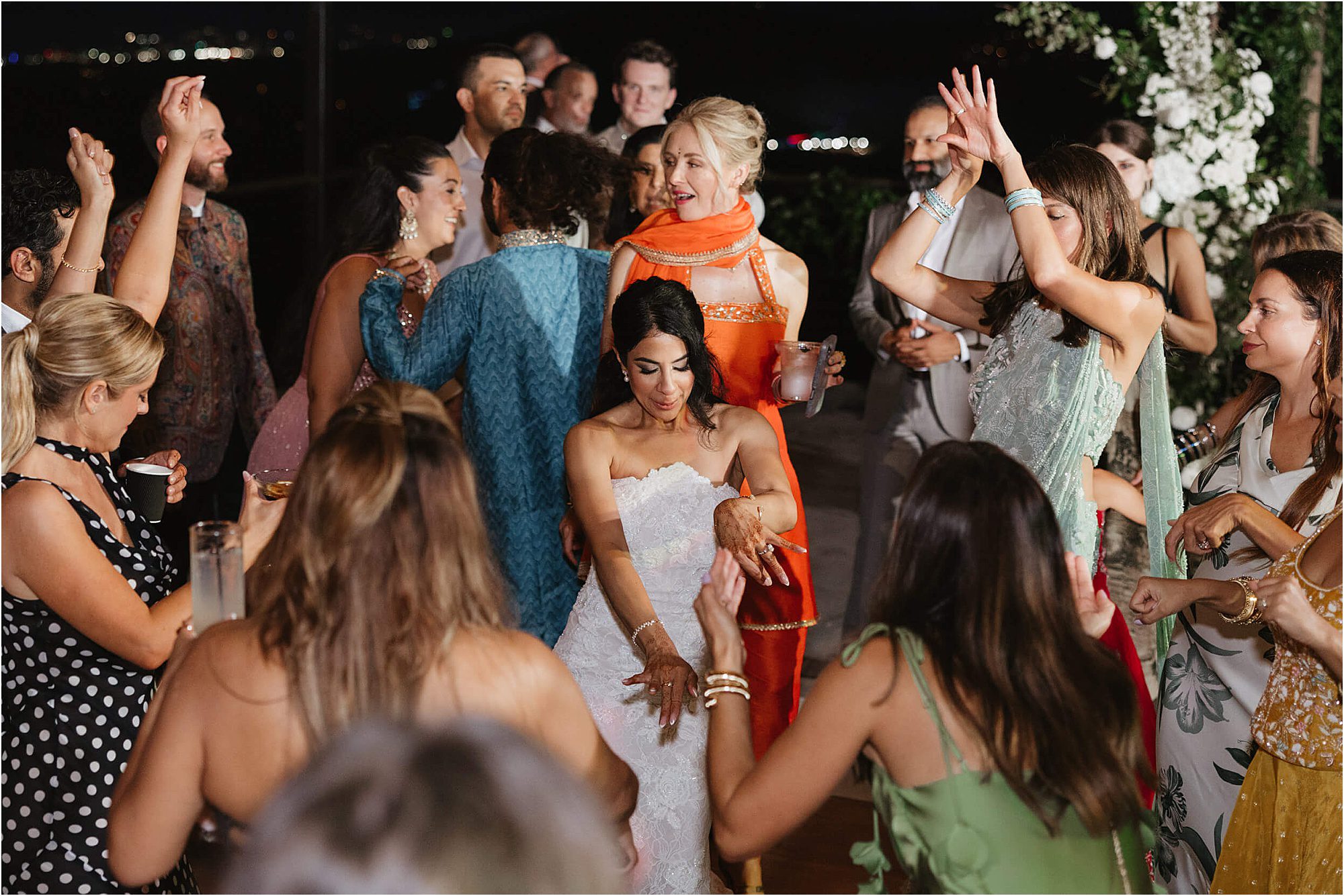 A bride in a white dress dances joyfully, surrounded by guests in colorful outfits with raised hands, celebrating together at a lively nighttime wedding reception—an atmosphere created with the help of expert wedding advice from wedding DJs.