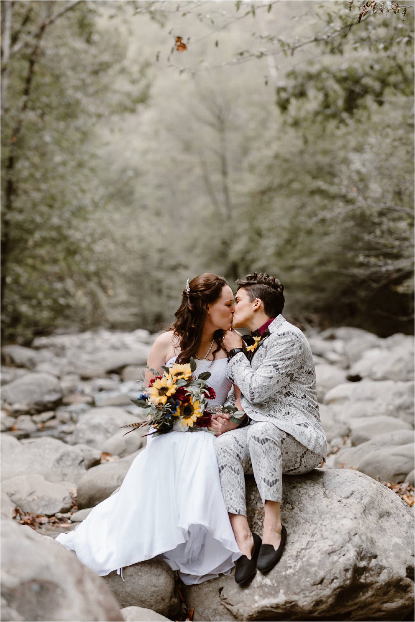 bride and bride kiss while sitting on riverbed