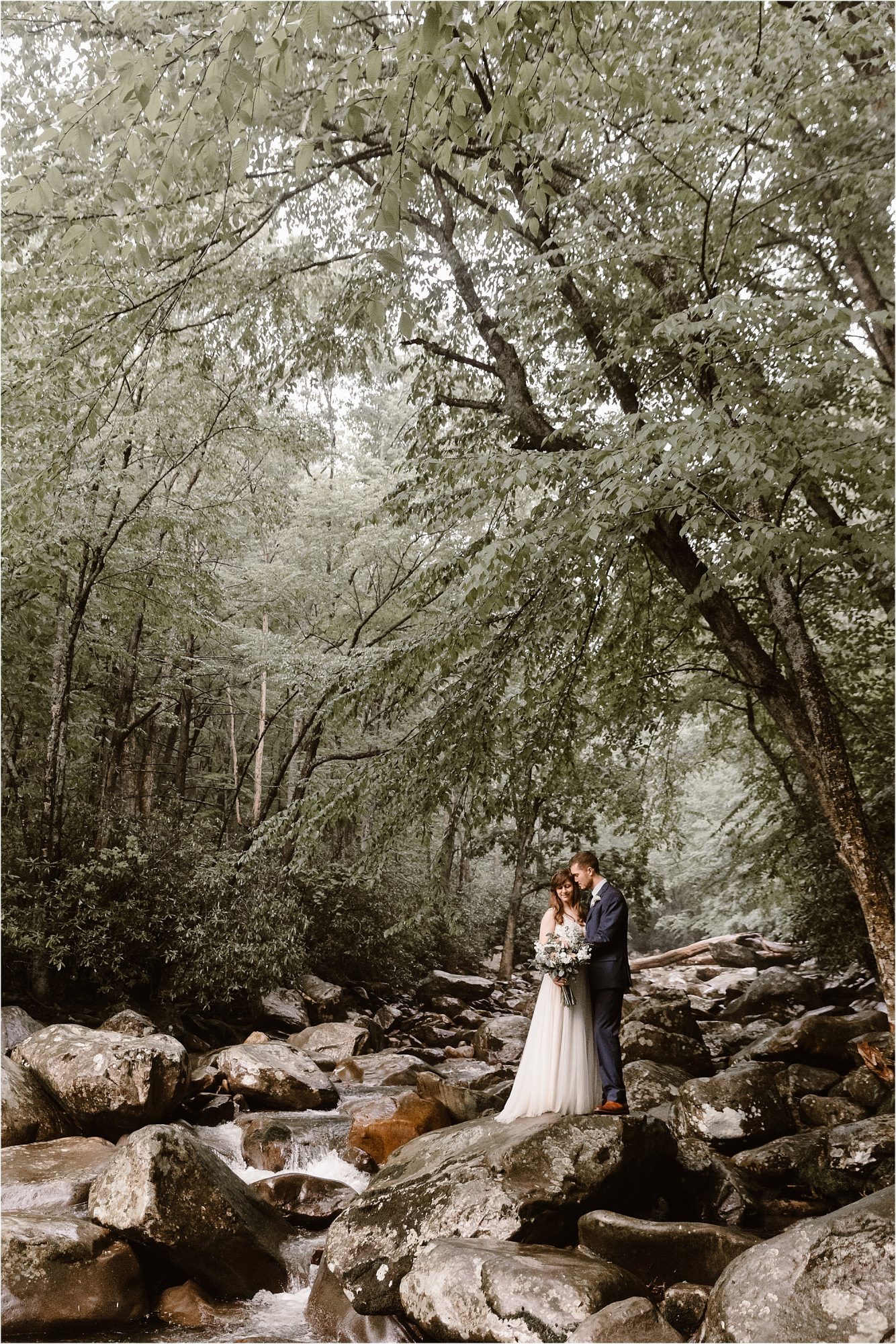 bride and groom embrace while standing on large rocks on bank of river