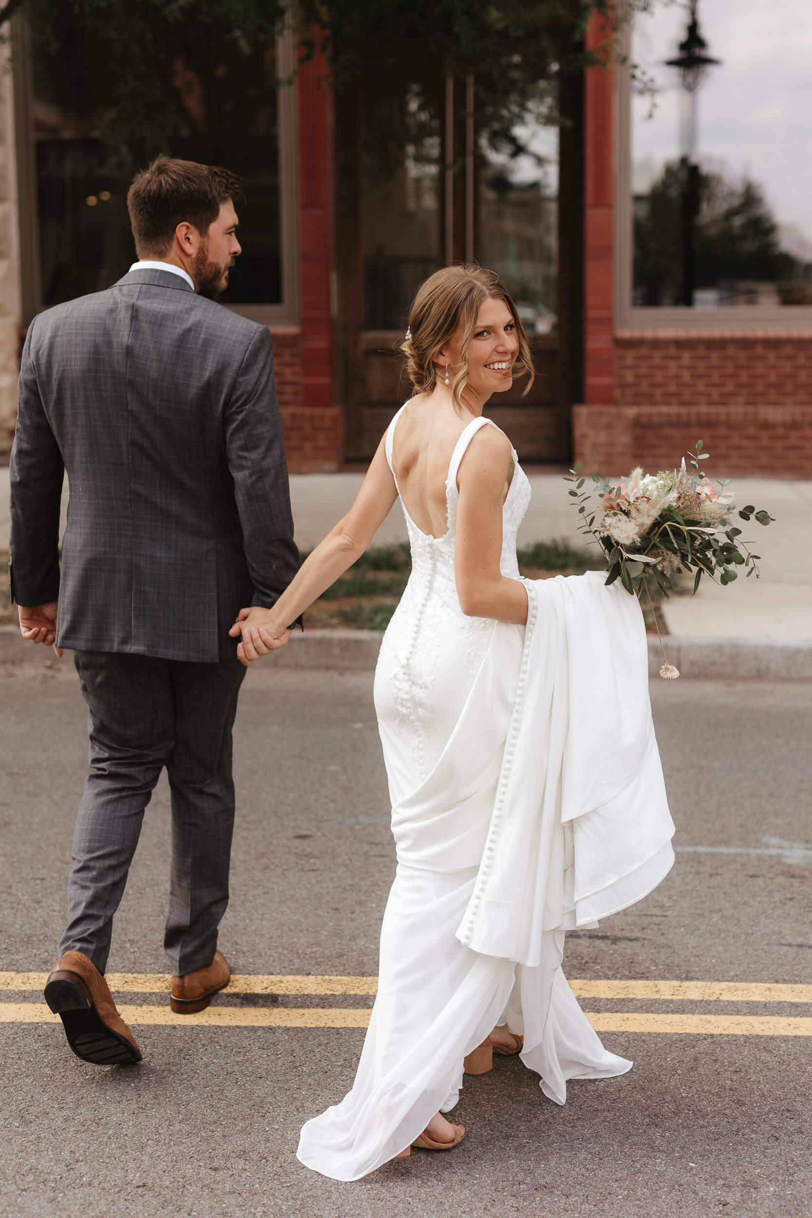 A bride and groom walk hand in hand down a street. The bride wears a white wedding dress and holds a bouquet, smiling back over her shoulder. The groom is in a dark suit. They walk on a double yellow line with buildings in the background.