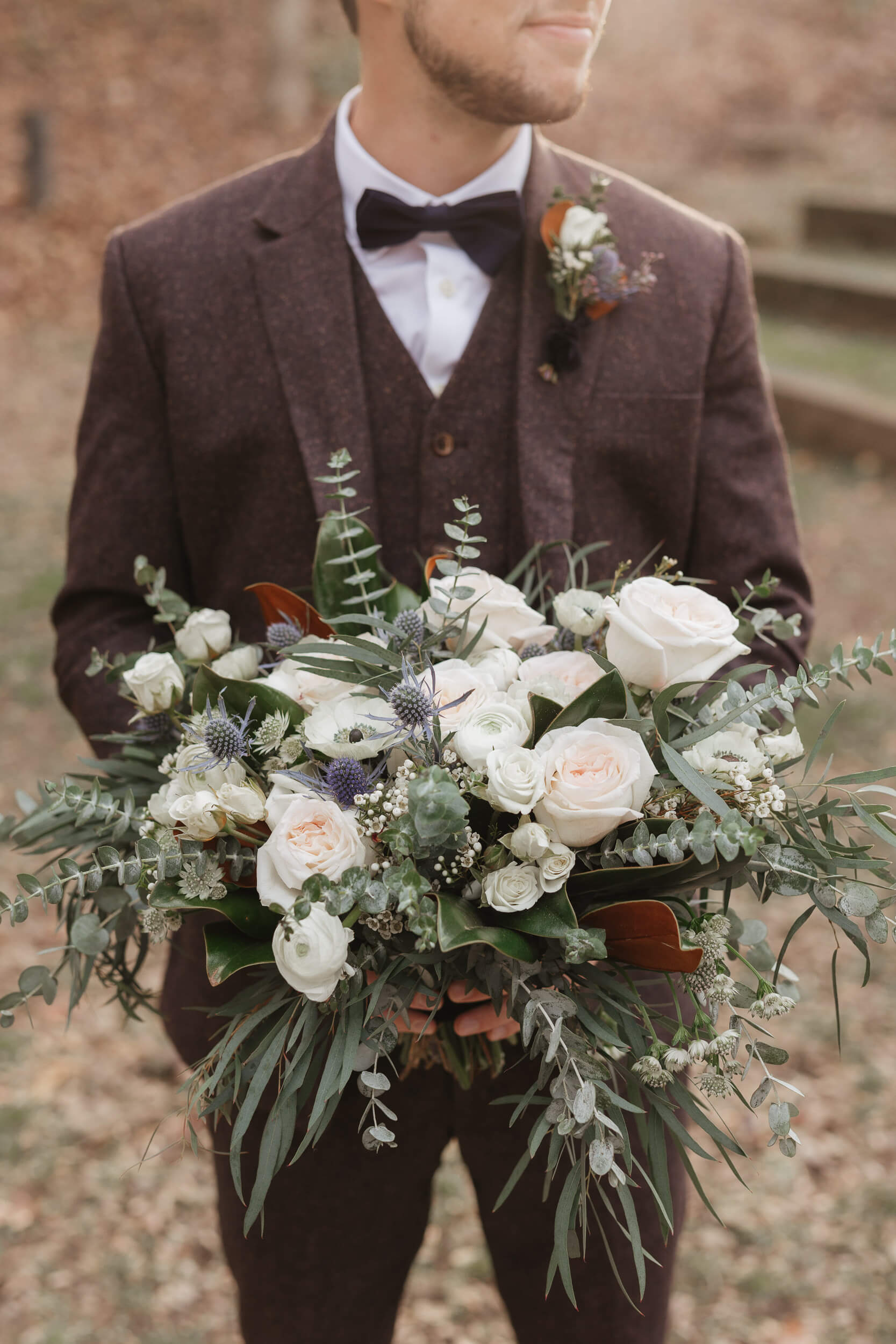 A person in a dark suit with a bow tie holds a large bouquet of white roses, eucalyptus leaves, and assorted greenery. They stand outdoors in an area with fallen leaves and steps in the background.