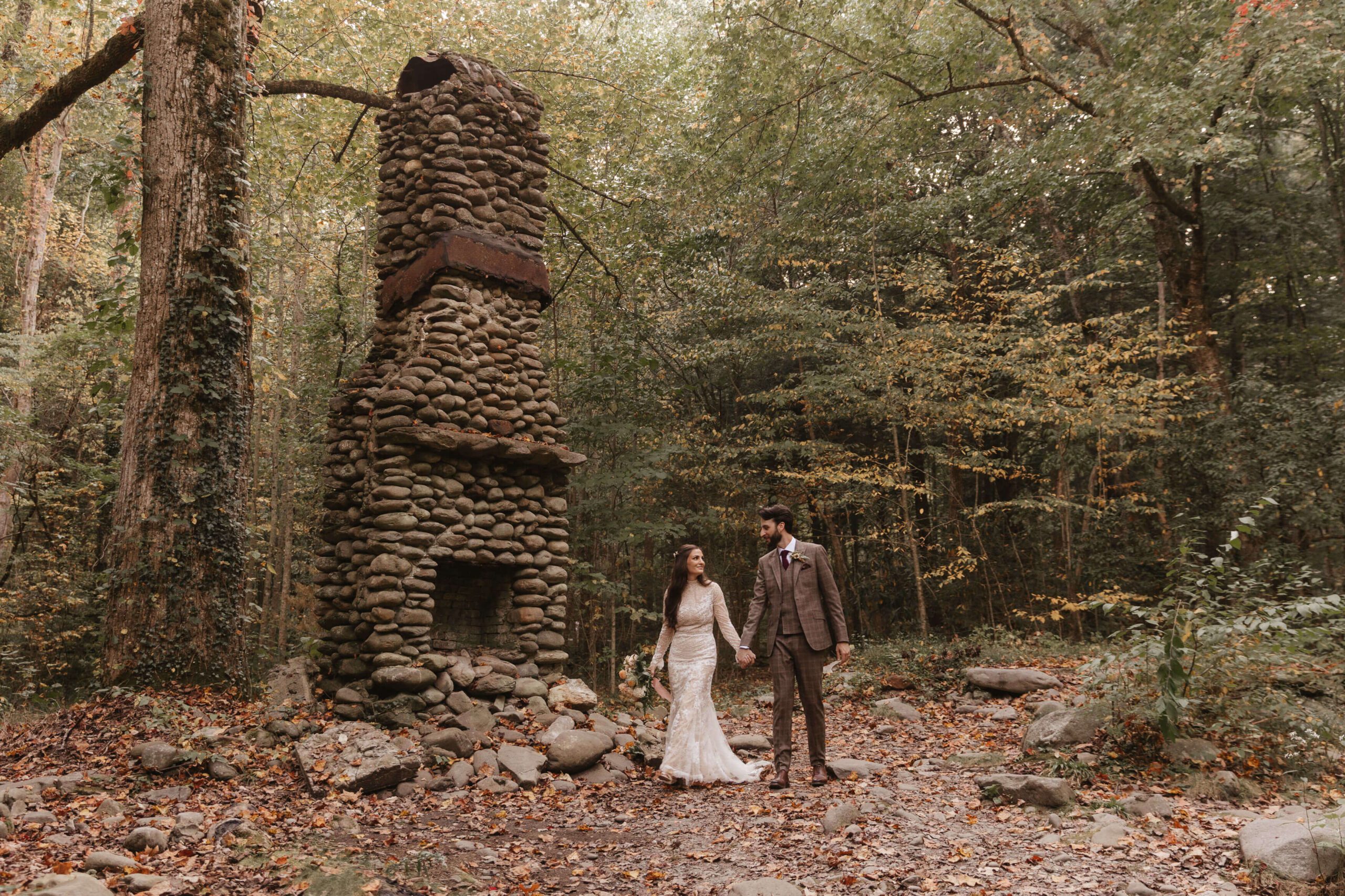 A bride and groom stand hand in hand in a forest clearing. Behind them is a tall, stone chimney structure. The forest is lush with green trees, and the ground is covered in fallen leaves. The scene feels rustic and serene.