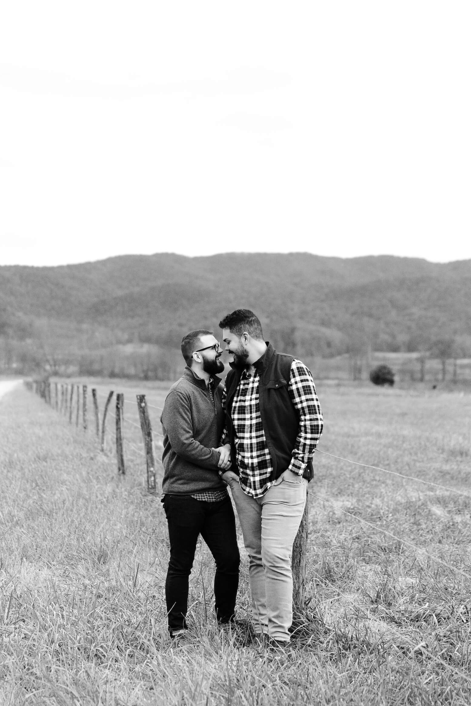 black and white engagement photo of two men leaning on fence post in the mountains