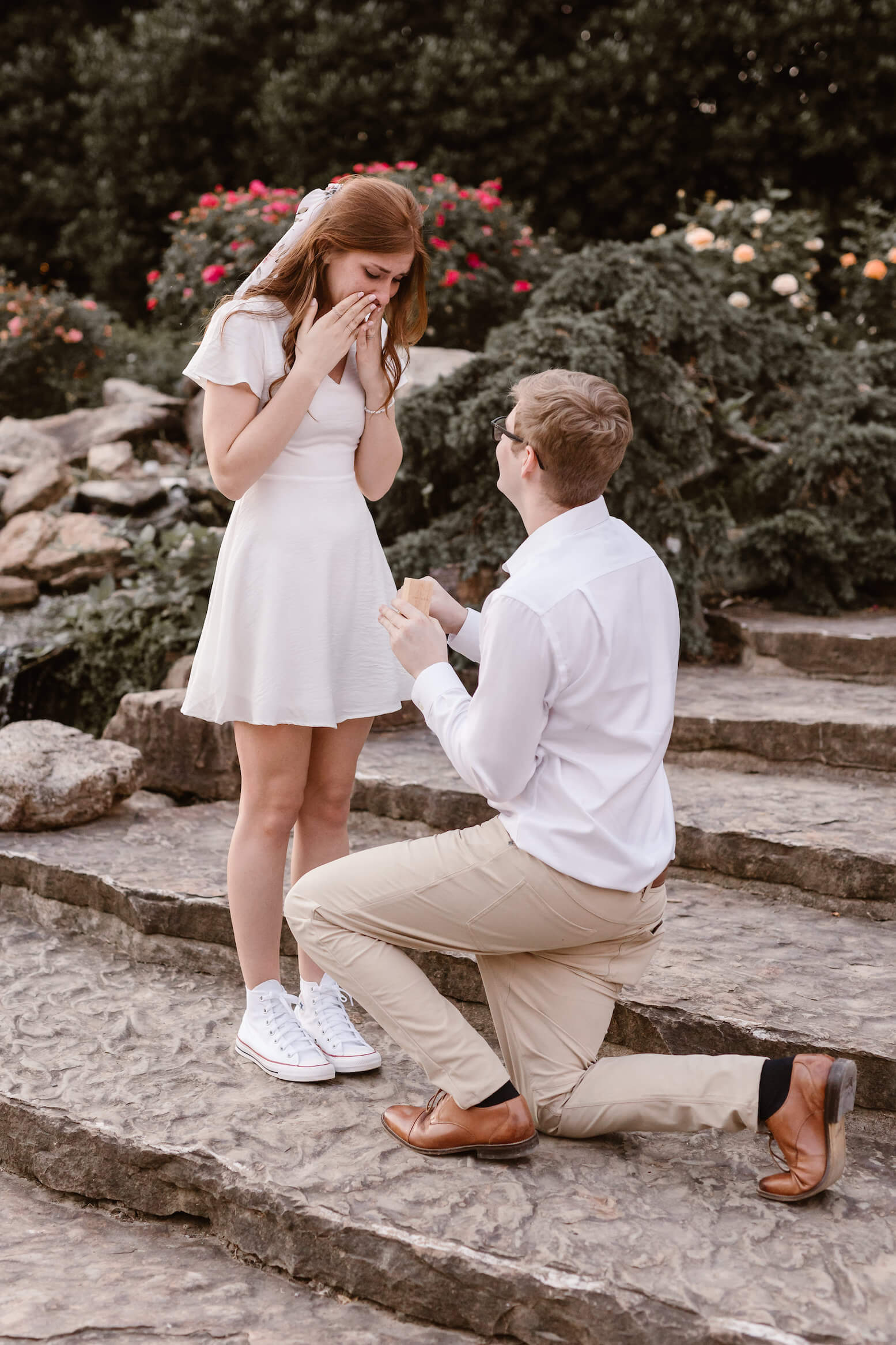 man proposing to woman in short white dress while holding ring box on one knee