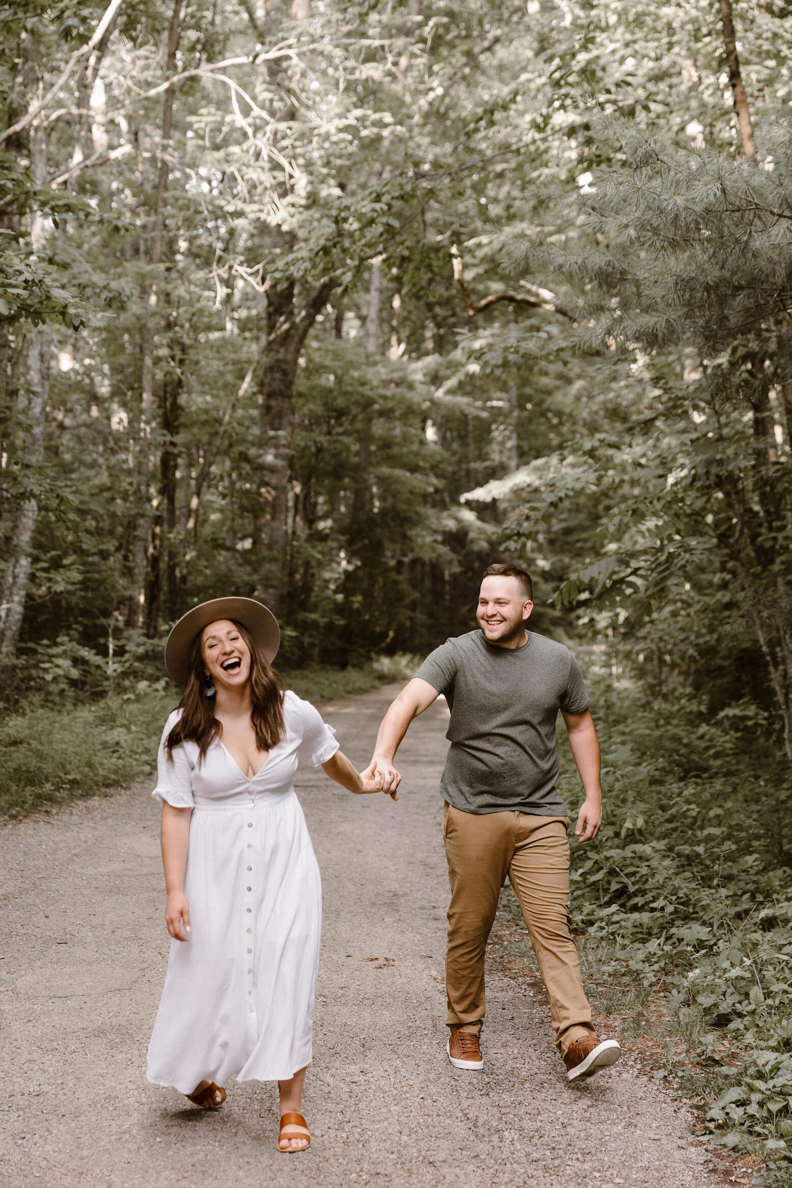 man and woman in brown hat laughing while walking during engagement session in the forest