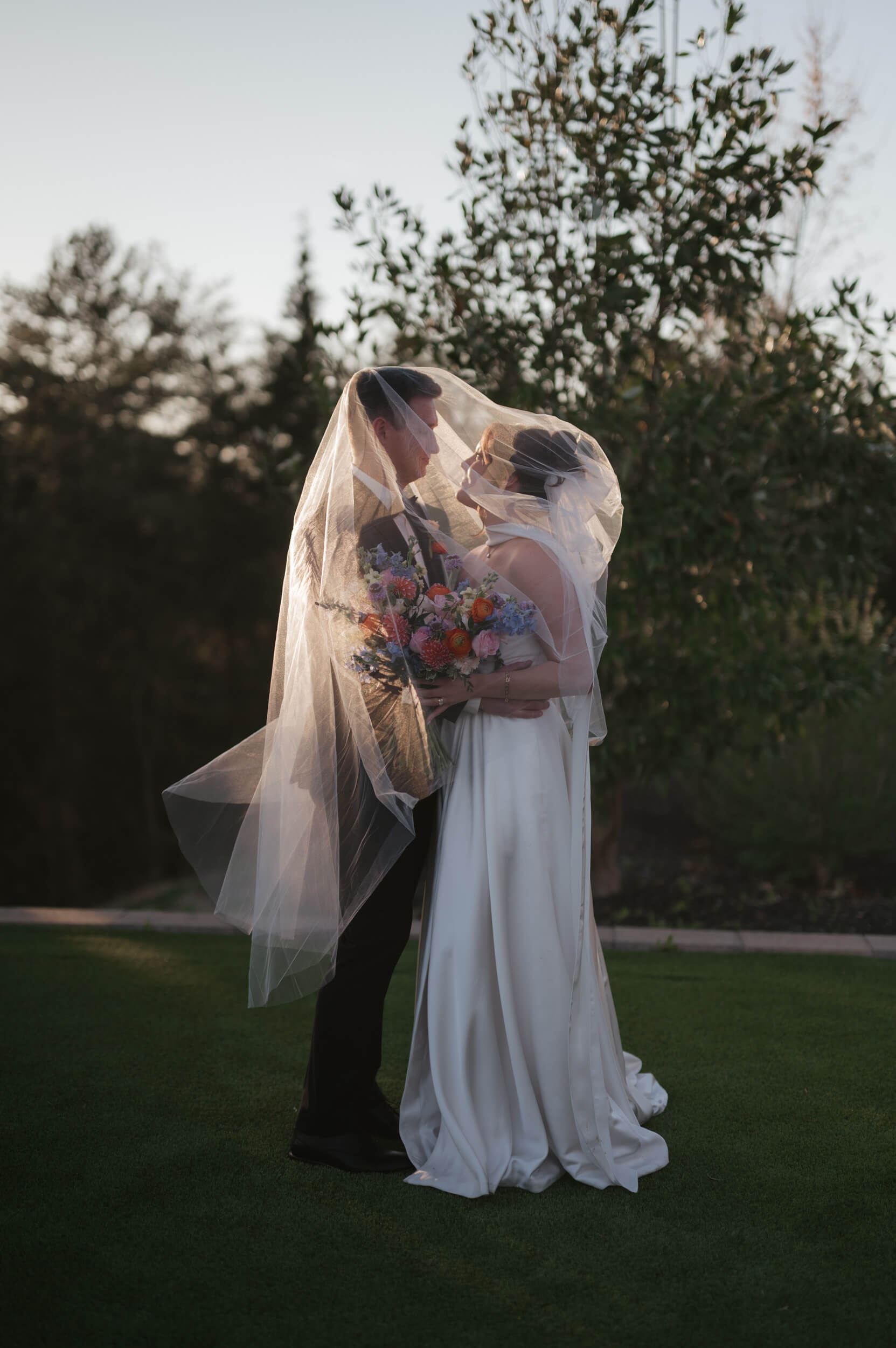 A bride and groom stand closely together outdoors under a sheer veil, holding a colorful bouquet and gazing into each other’s eyes, surrounded by greenery at sunset.
