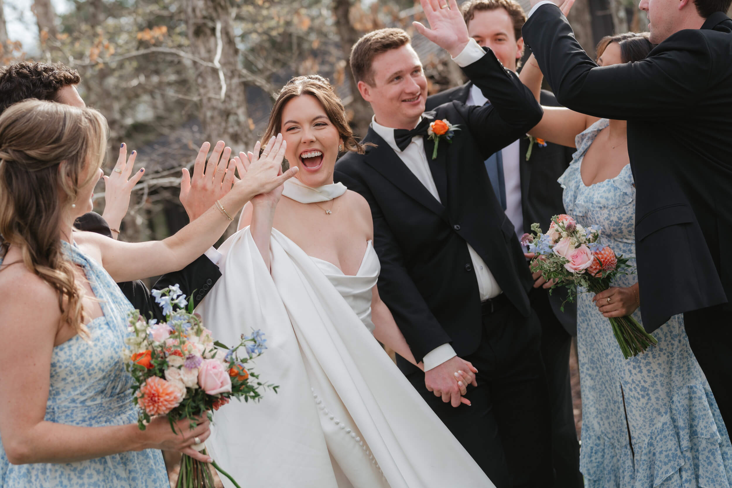A joyful bride and groom hold hands and high-five friends in floral dresses and suits, surrounded by bouquets, celebrating outdoors on their wedding day.