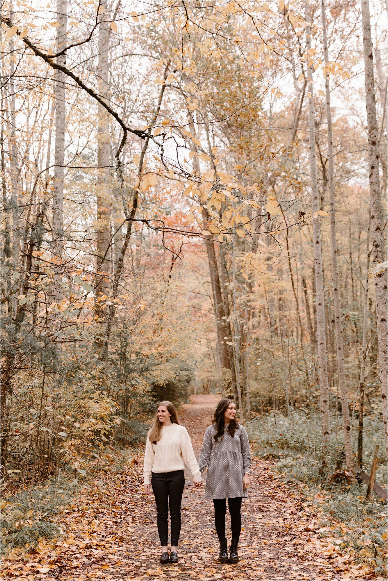 two women holding hands and walking down forest path at autumn engagement session