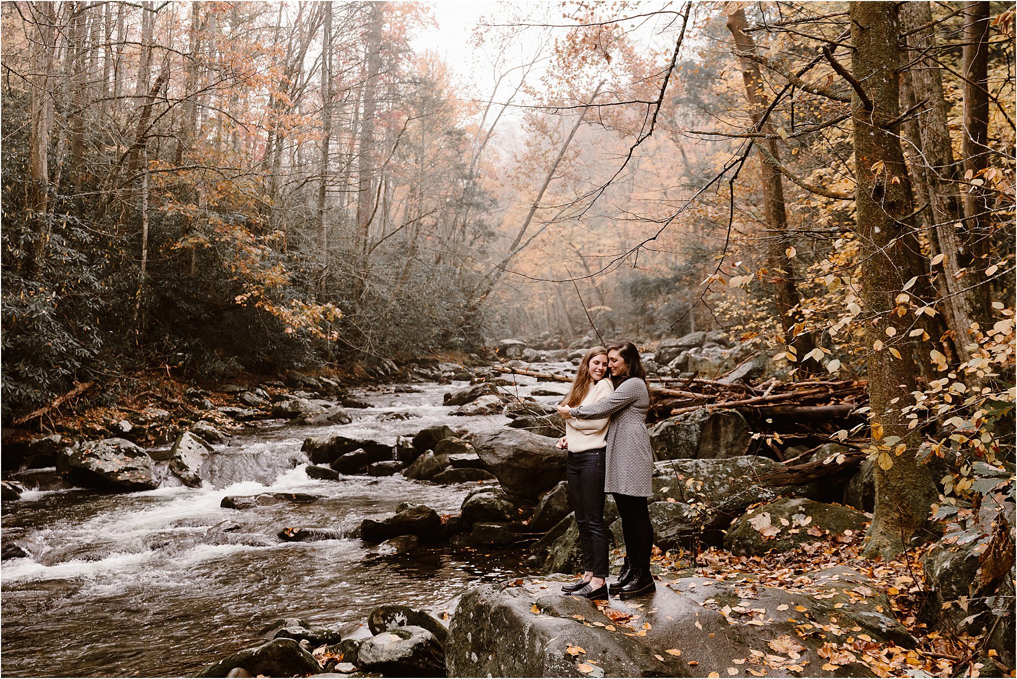 two women hugging on large rock by flowing river in the Fall