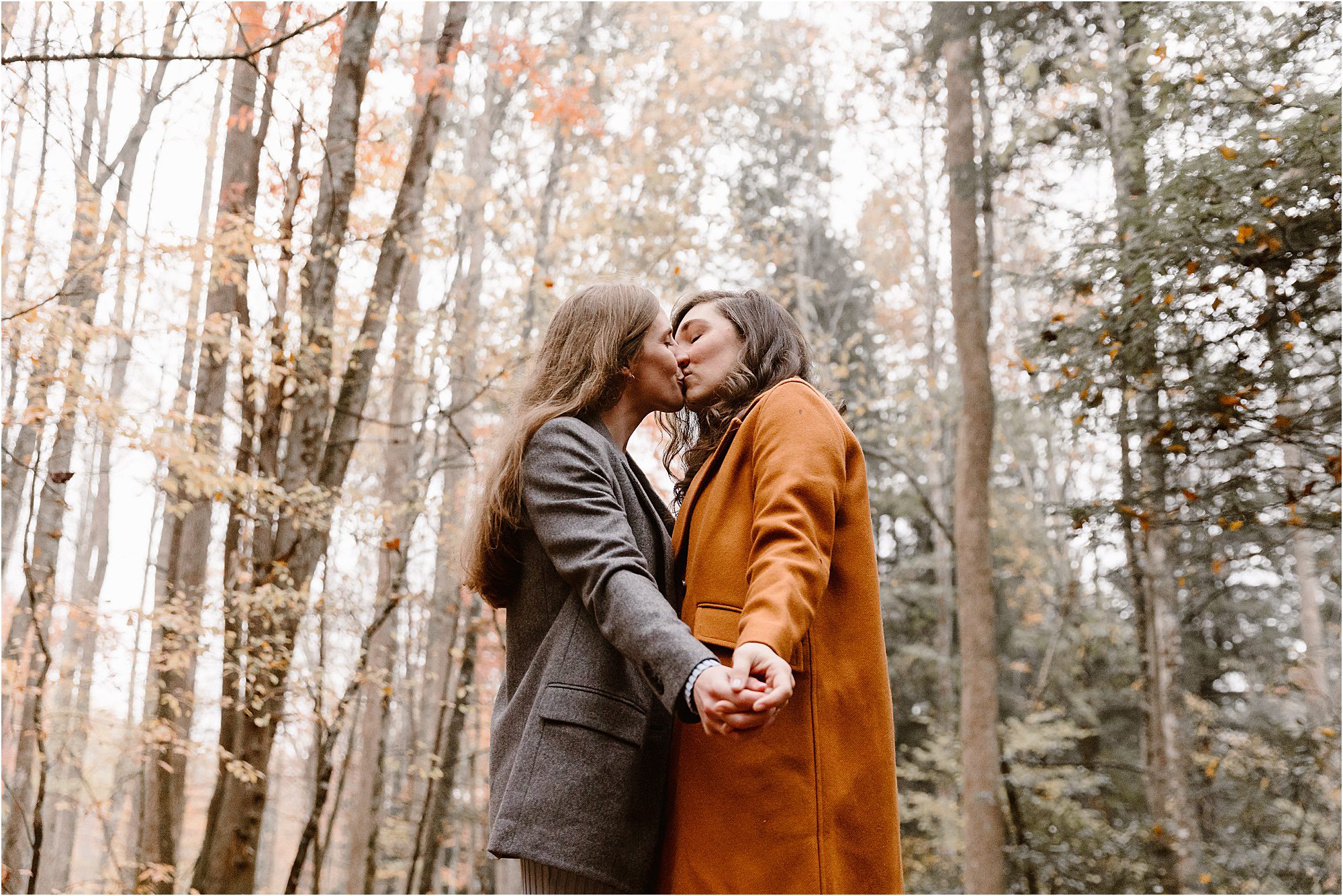 two women kissing and holding hands with forest trees in the background