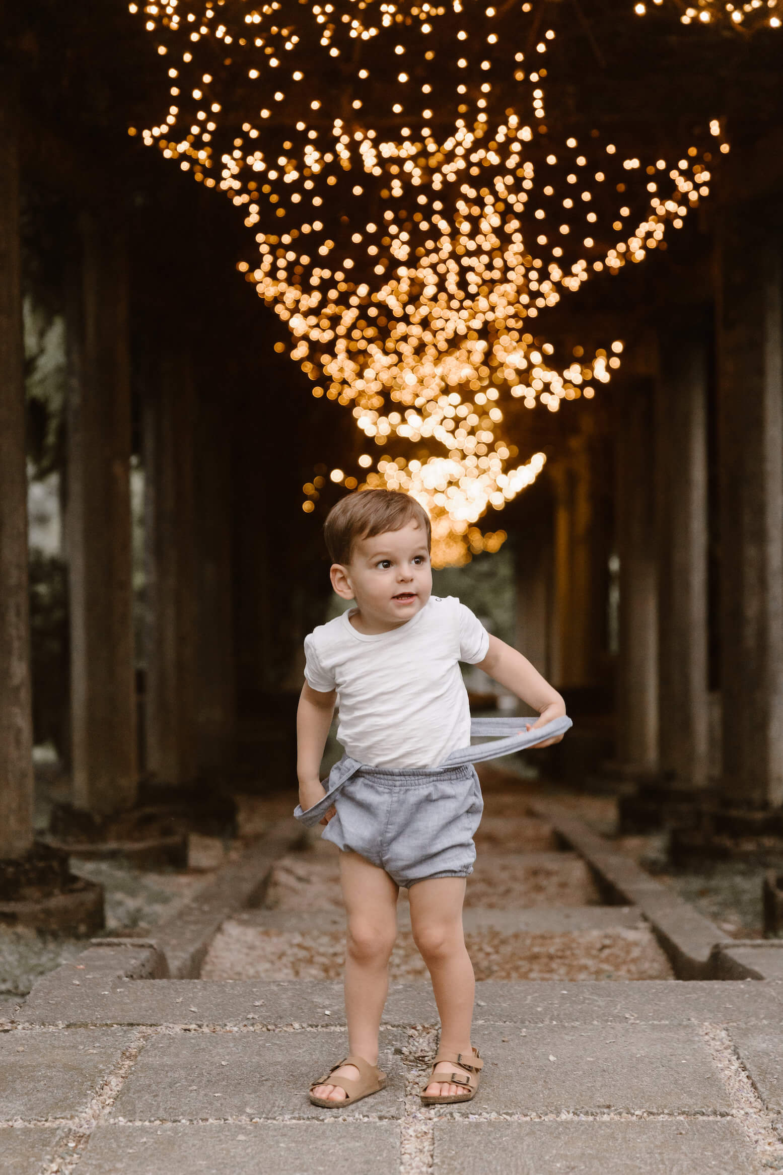 little boy holding suspenders while standing in front of stairs with lights during family photography session with Erin Morrison