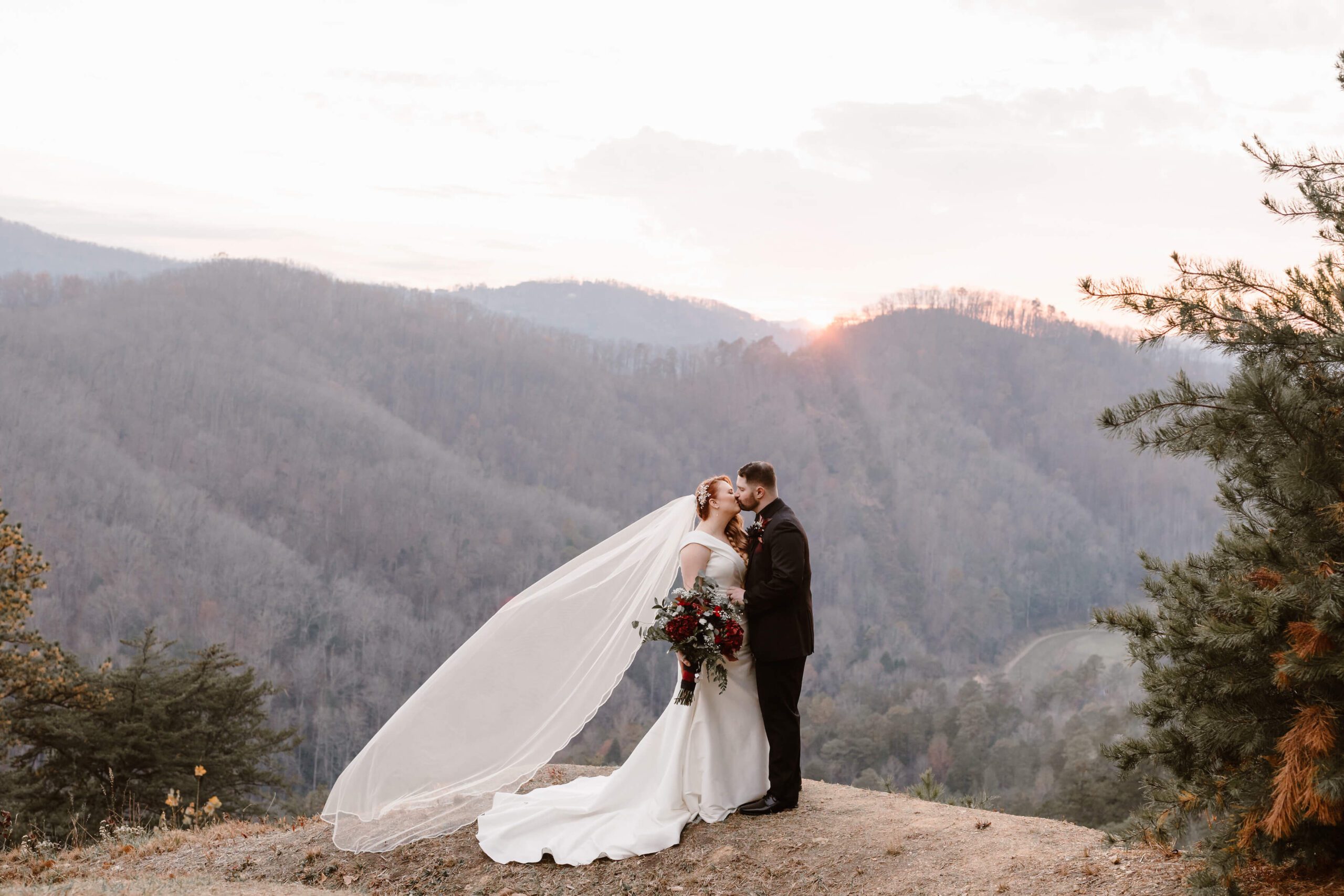 bride in long veil holding red bouquet while kissing man in black suit standing in mountains