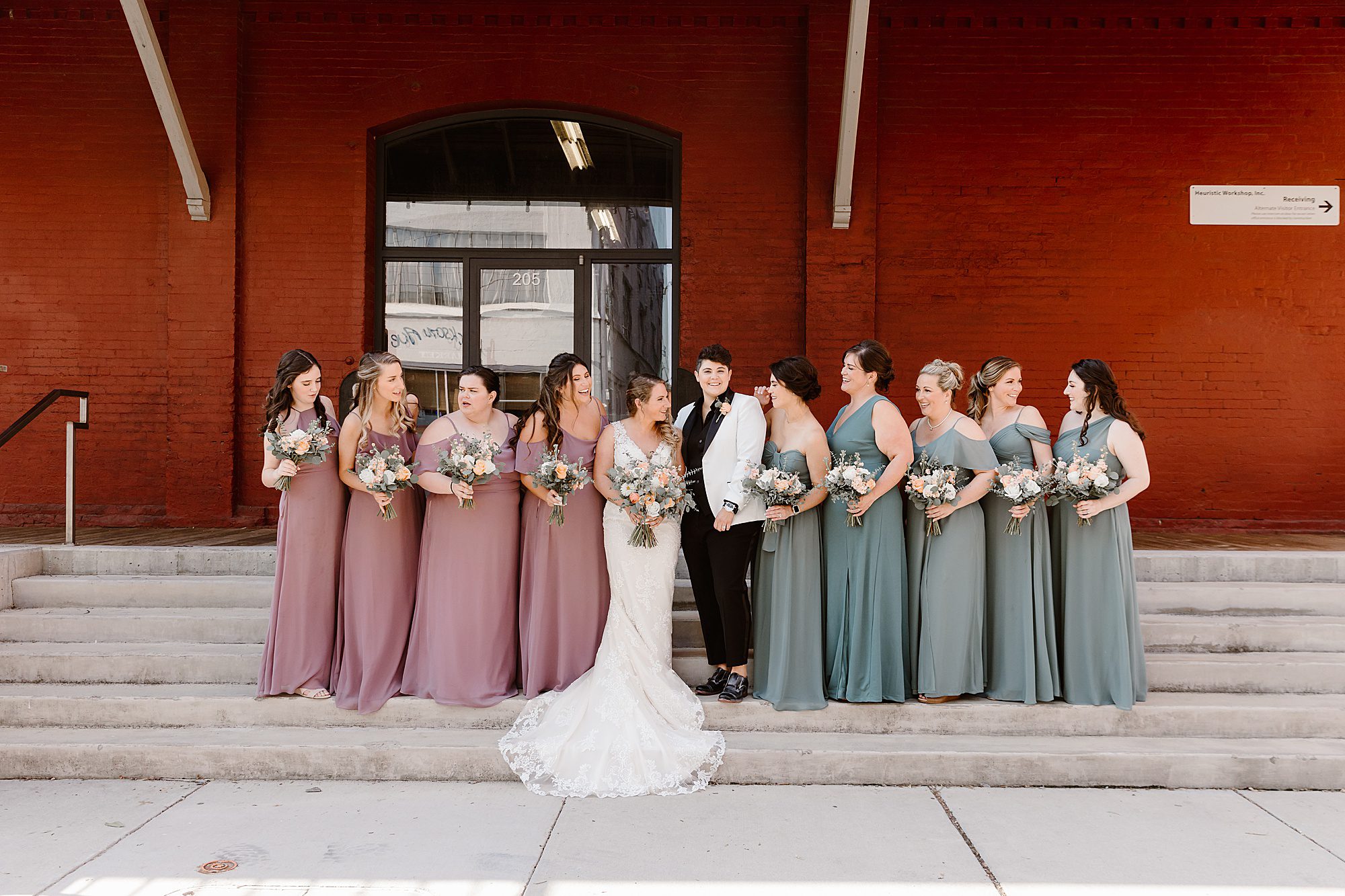 bride and bride stand with wedding party on stairs in front of red wall