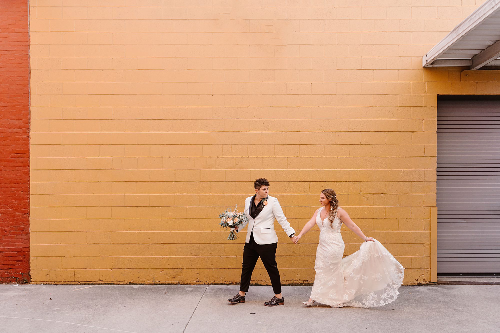 bride in black and white suit leads bride in white wedding dress across yellow wall