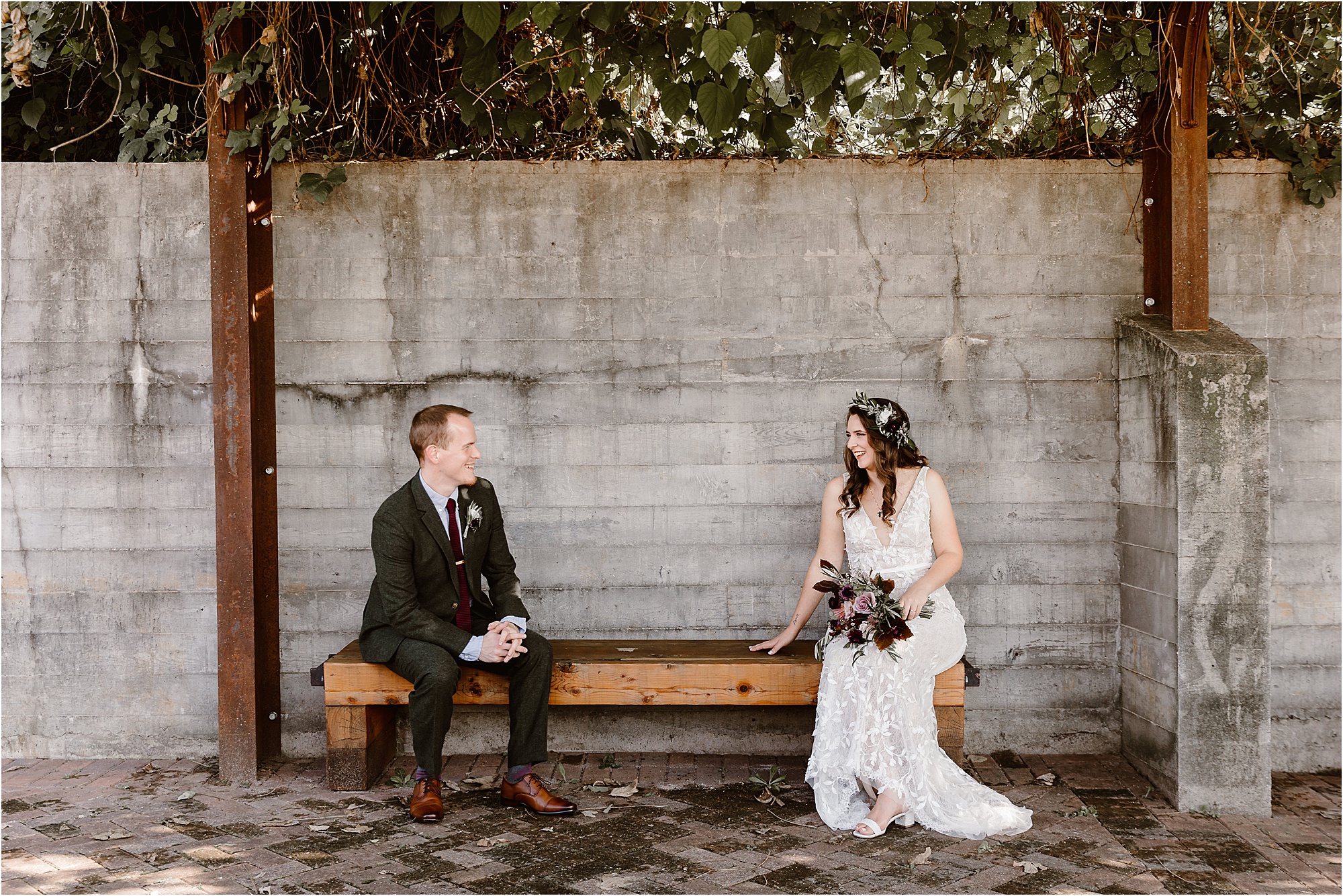 man in tweed jacket sits on bench opposite side of woman in sleeveless white wedding dress wearing flower crown