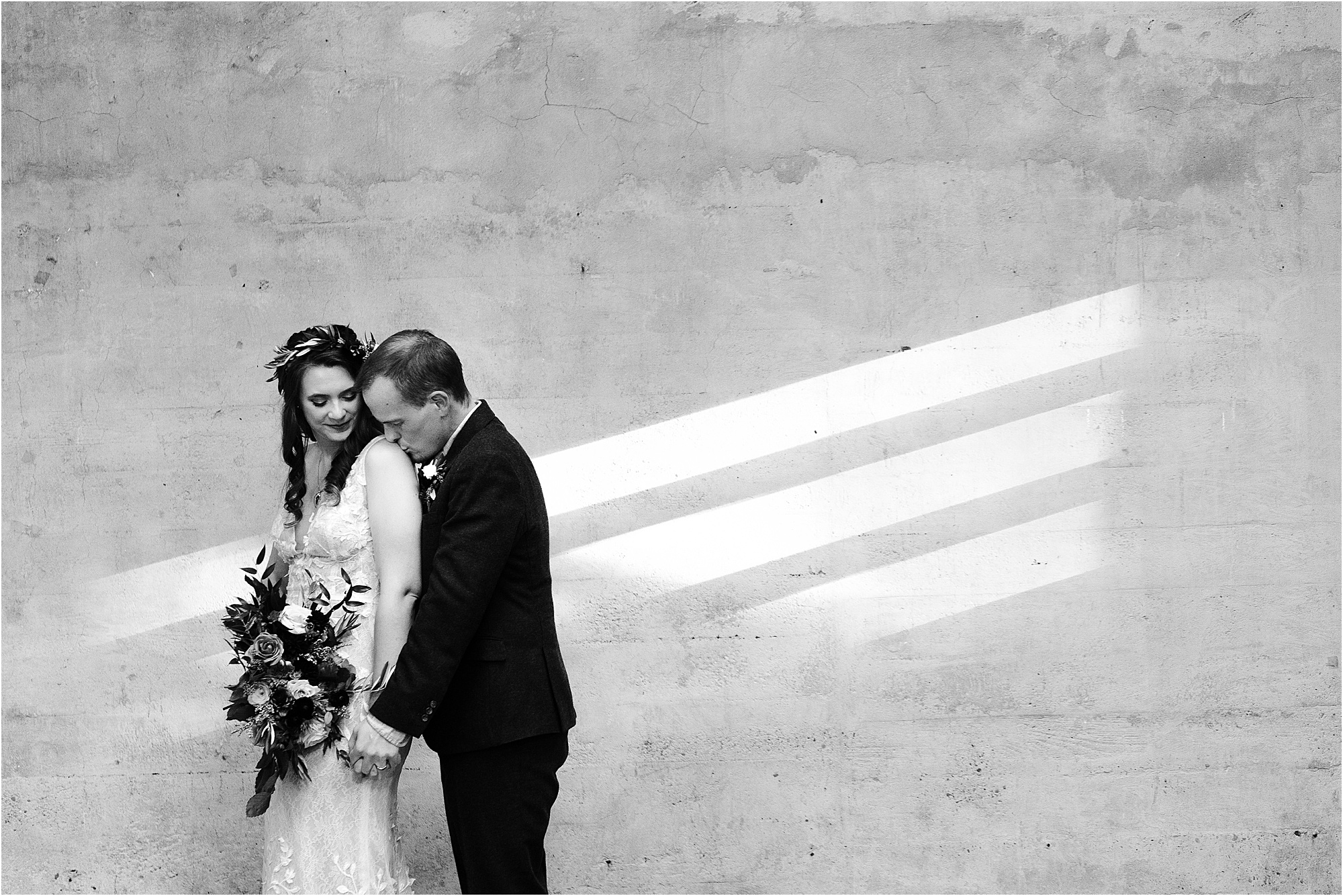 groom kisses bride on shoulder along wall with direct lighting