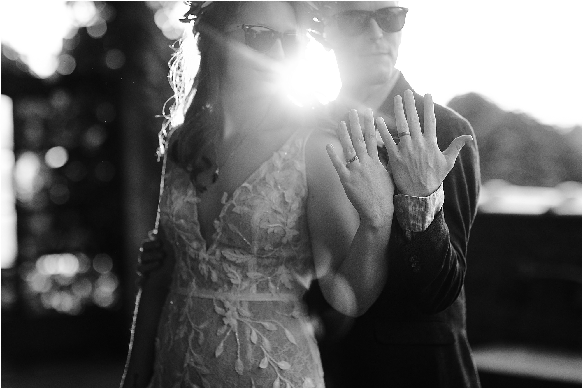 black and white photo of bride and groom holding up rings while wearing sunglasses