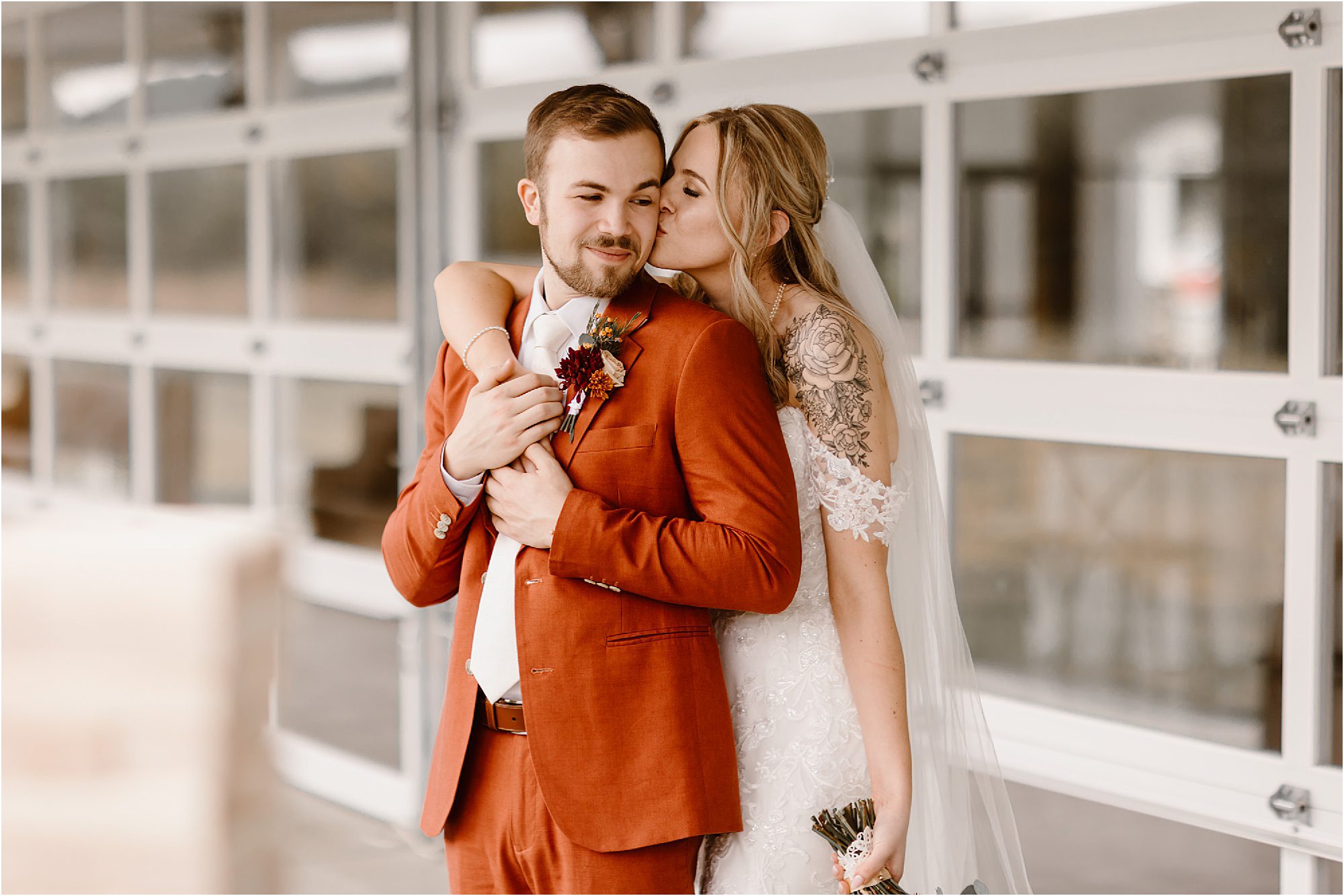 bride hugging and kissing groom in burnt orange suit on modern patio