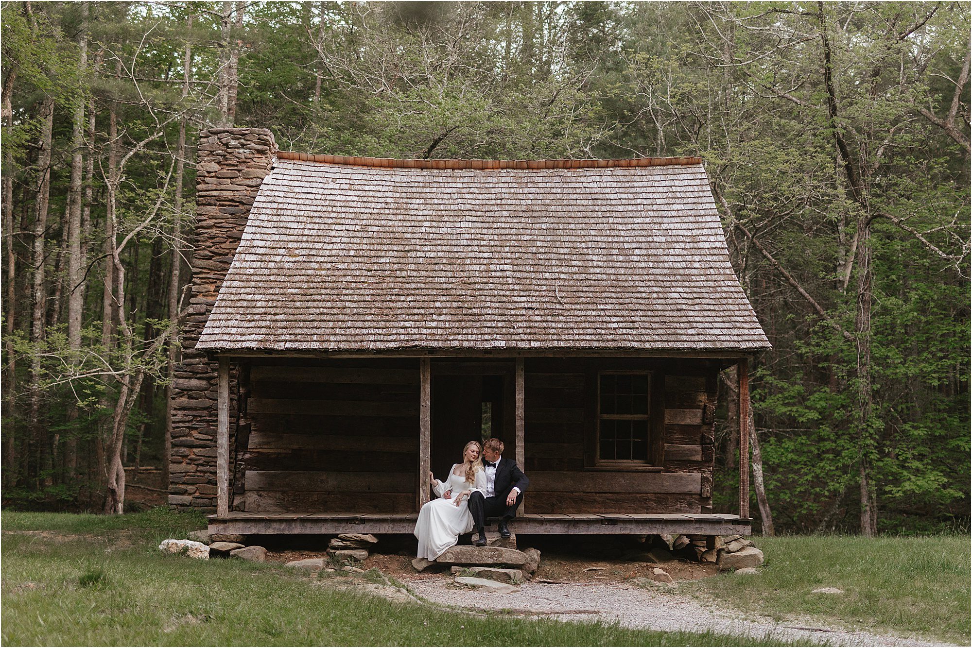 A couple dressed in formal attire sits together on the porch of a rustic wooden cabin surrounded by trees and greenery, capturing the essence of a Great Smoky Mountain Elopement.