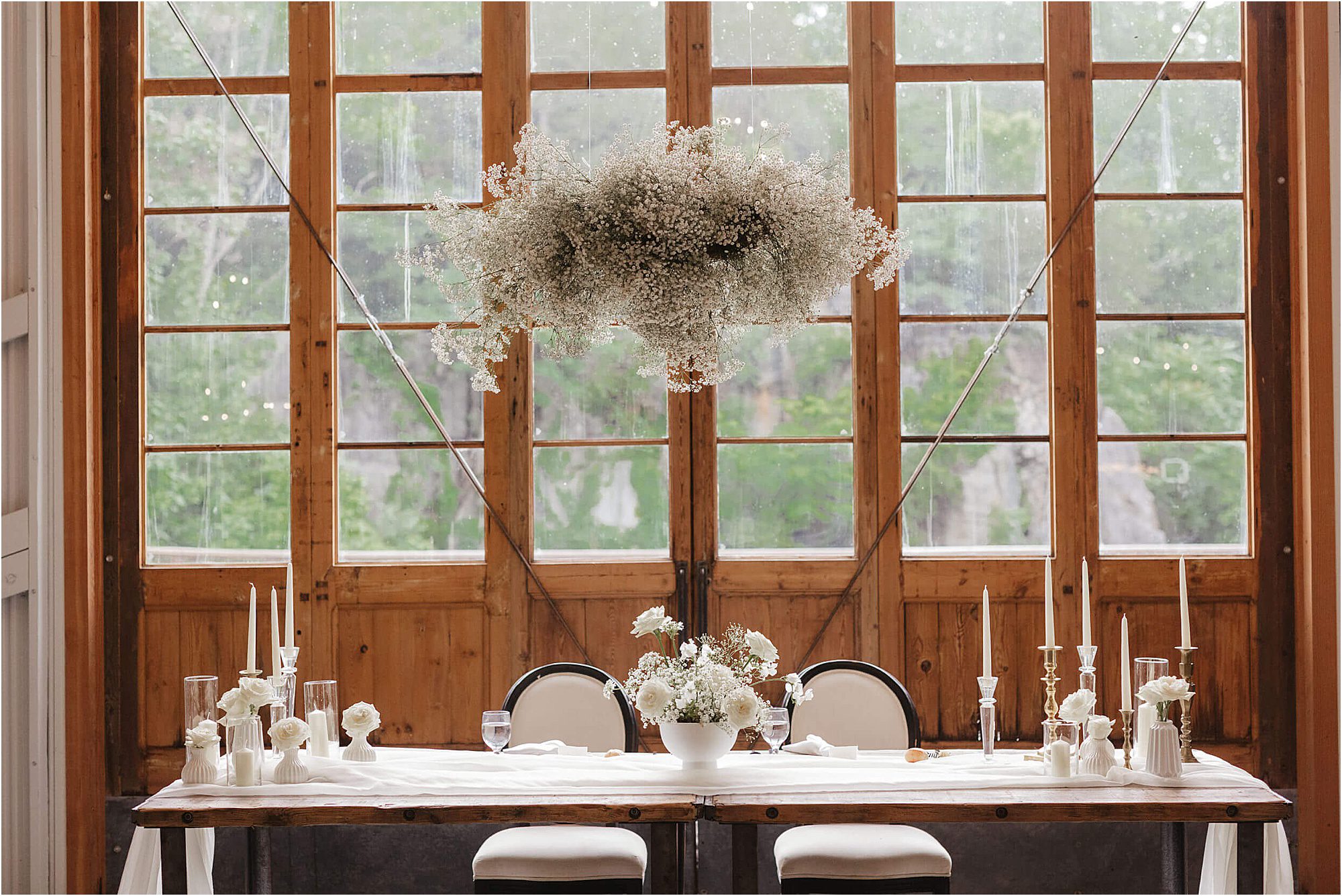 A wooden table set for two with white flowers, candles, and glassware sits in front of tall windows. A Baby’s Breath Wedding floral arrangement hangs gracefully above the table. Natural light fills the elegant, rustic room.