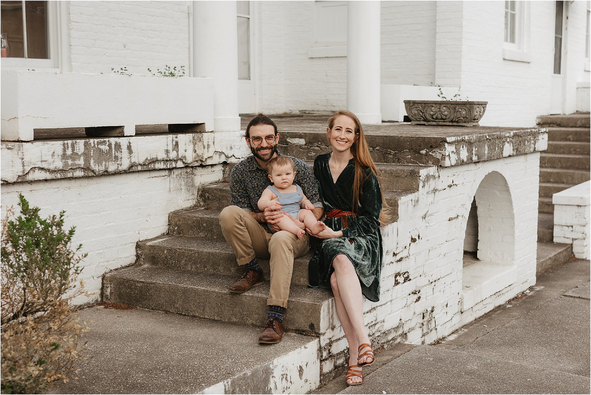 A family sits on outdoor steps, radiating warmth amidst the backdrop of a white brick building. Two adults smile brightly, dressed in casual attire—a dark dress and a patterned shirt with khakis—while cradling a baby in light clothing. It’s a timeless moment reminiscent of classic Bleak House Family Photos.