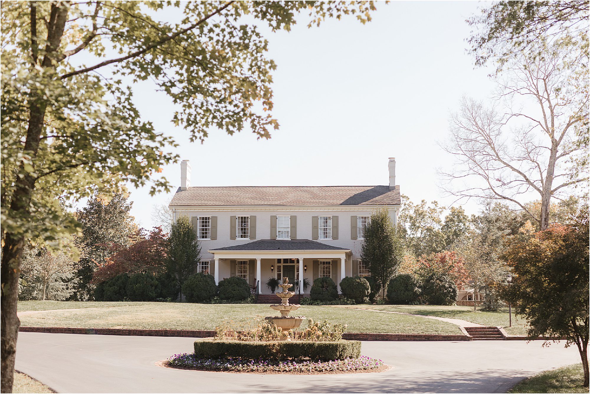 A large, elegant house with white columns and symmetrical windows is set back from a circular driveway, perfect for a Sunday wedding. A fountain surrounded by flowers is in the foreground, with trees and shrubs providing a scenic backdrop under a clear sky.