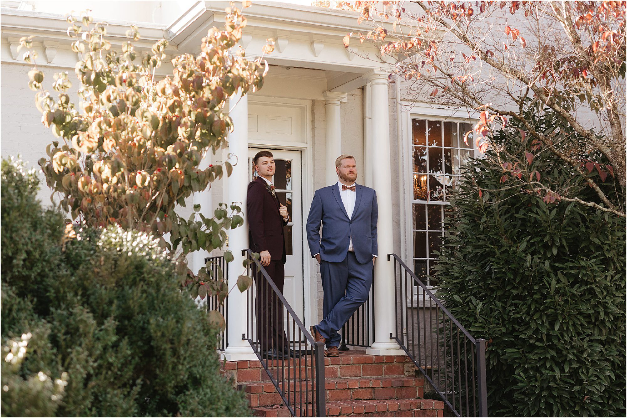 Two people in suits stand on a brick staircase outside a building with white columns, attending a Sunday wedding. One wears a dark suit, the other a blue suit. The building is surrounded by green and brown foliage, with a large window to the right.