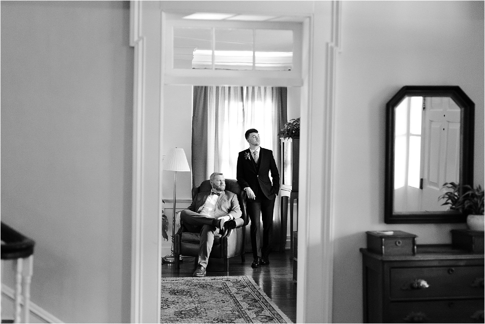 A black and white photo captures the timeless elegance of a Sunday wedding, with one man seated on a chair in light-colored attire, while the other stands beside him in a dark suit. The room features a patterned rug, soft lamp glow, elegant curtains, and a mirror atop the dresser.