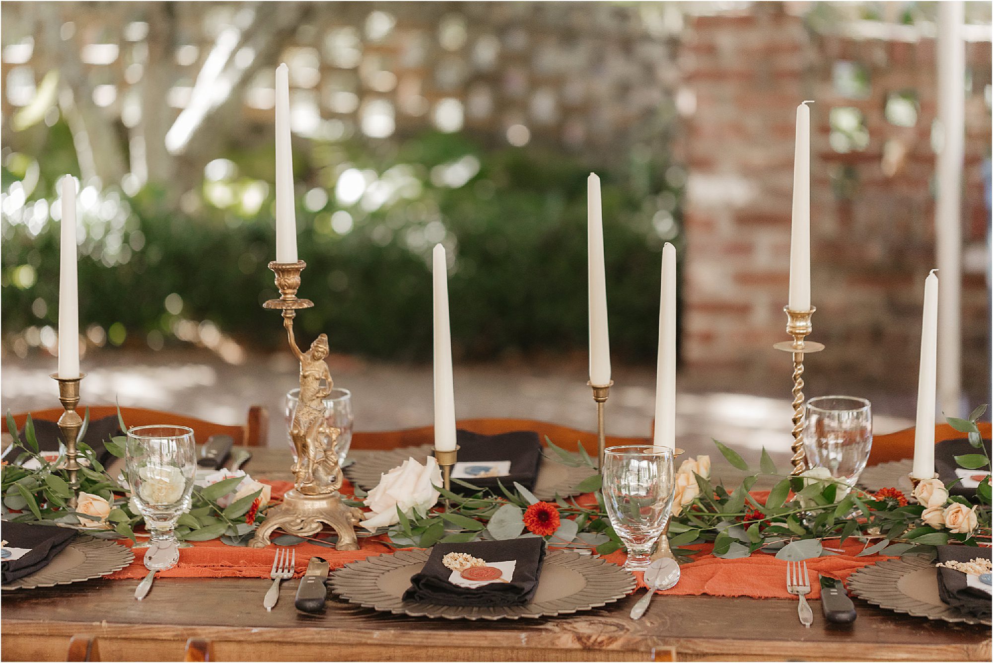 A beautifully set table outdoors for a Sunday wedding features tall white candles in ornate holders, greenery with small flowers as a centerpiece, black plates with gold accents, clear glassware, and silver cutlery. The background shows a blurred garden and brick wall.