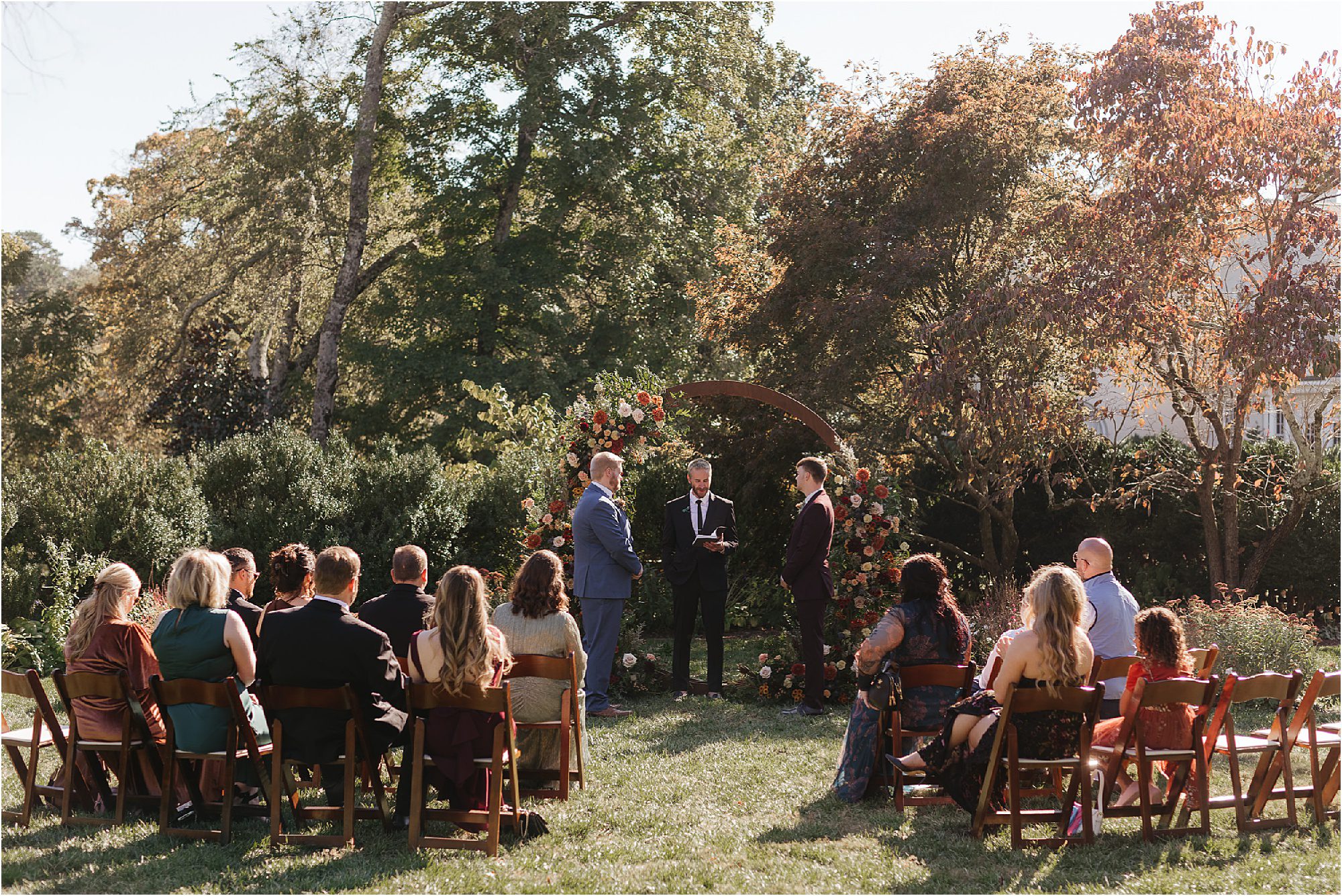A Sunday wedding unfolds outdoors in a garden setting with lush greenery. Two people stand at the altar adorned with a floral arch, while guests sit on wooden chairs facing the couple. The sky is clear and the sun is shining brightly.