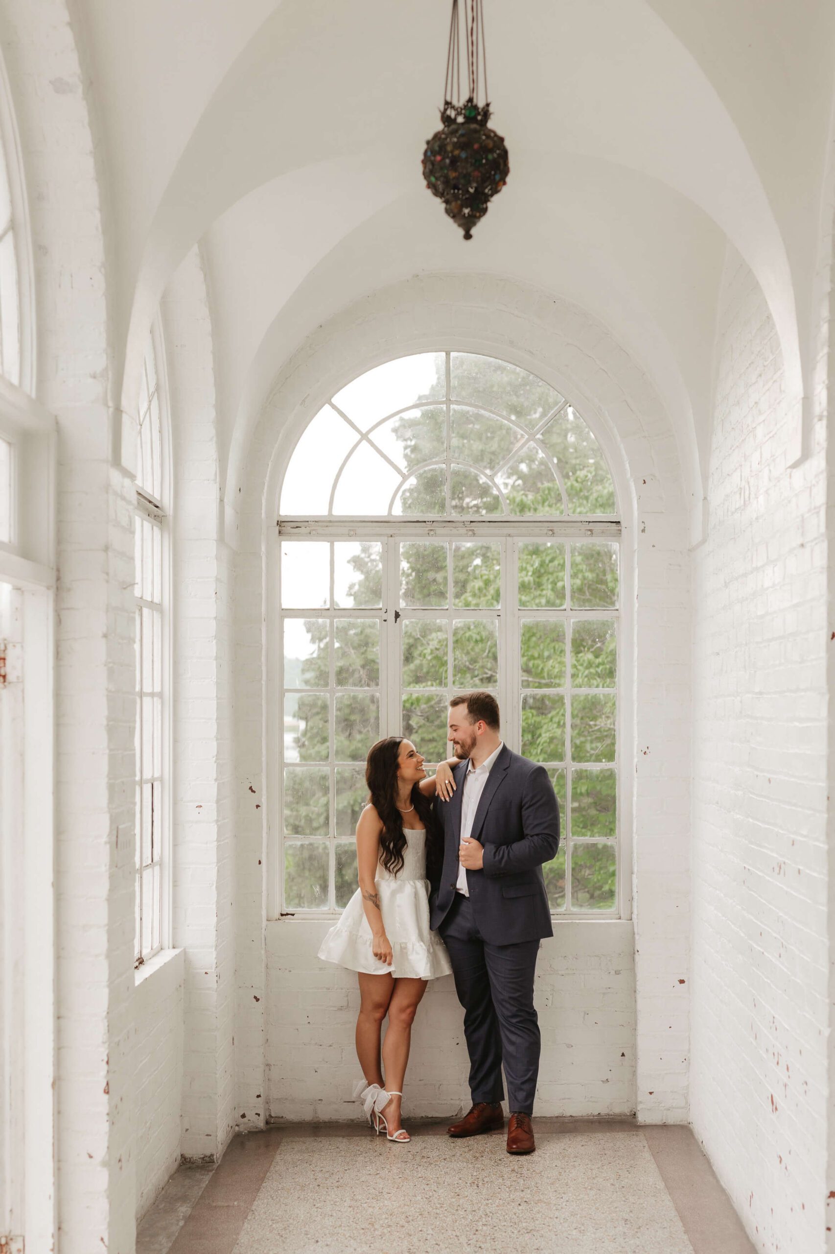 A couple stands in a hallway by large arched windows. The woman in a white dress gazes at the man in a dark suit, who leans against the wall. They share a joyful moment, surrounded by natural light.