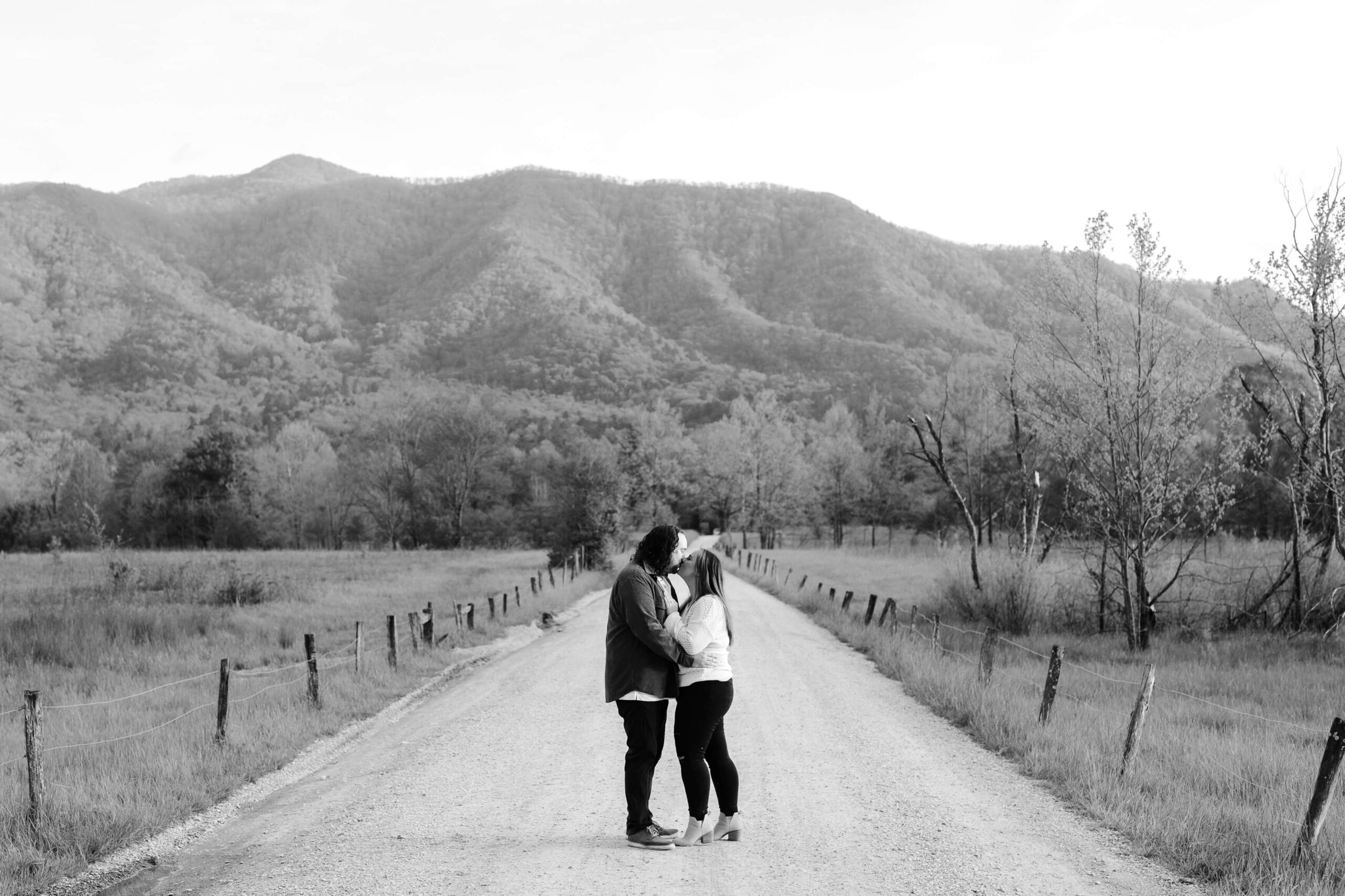 A couple stands in the middle of a dirt road, embracing, surrounded by open fields and mountains. The landscape is tranquil and partly wooded under a clear sky, in a black and white photograph.