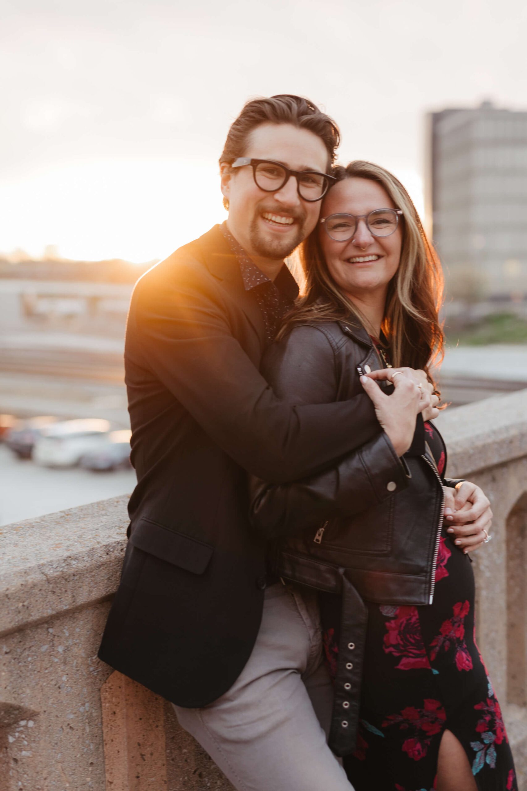 A couple smiling and hugging while leaning on a concrete railing. The setting sun casts a warm glow in the background. Both are wearing glasses and casual attire, with the woman in a leather jacket and the man in a blazer.