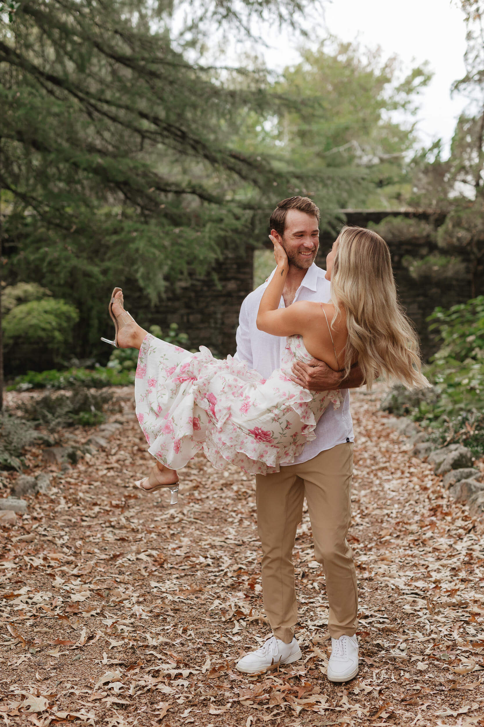 A man joyfully lifts a woman in a floral dress as they share a moment in a leafy, outdoor setting. The ground is covered with fallen leaves, and trees surround them. Both are smiling happily at each other.