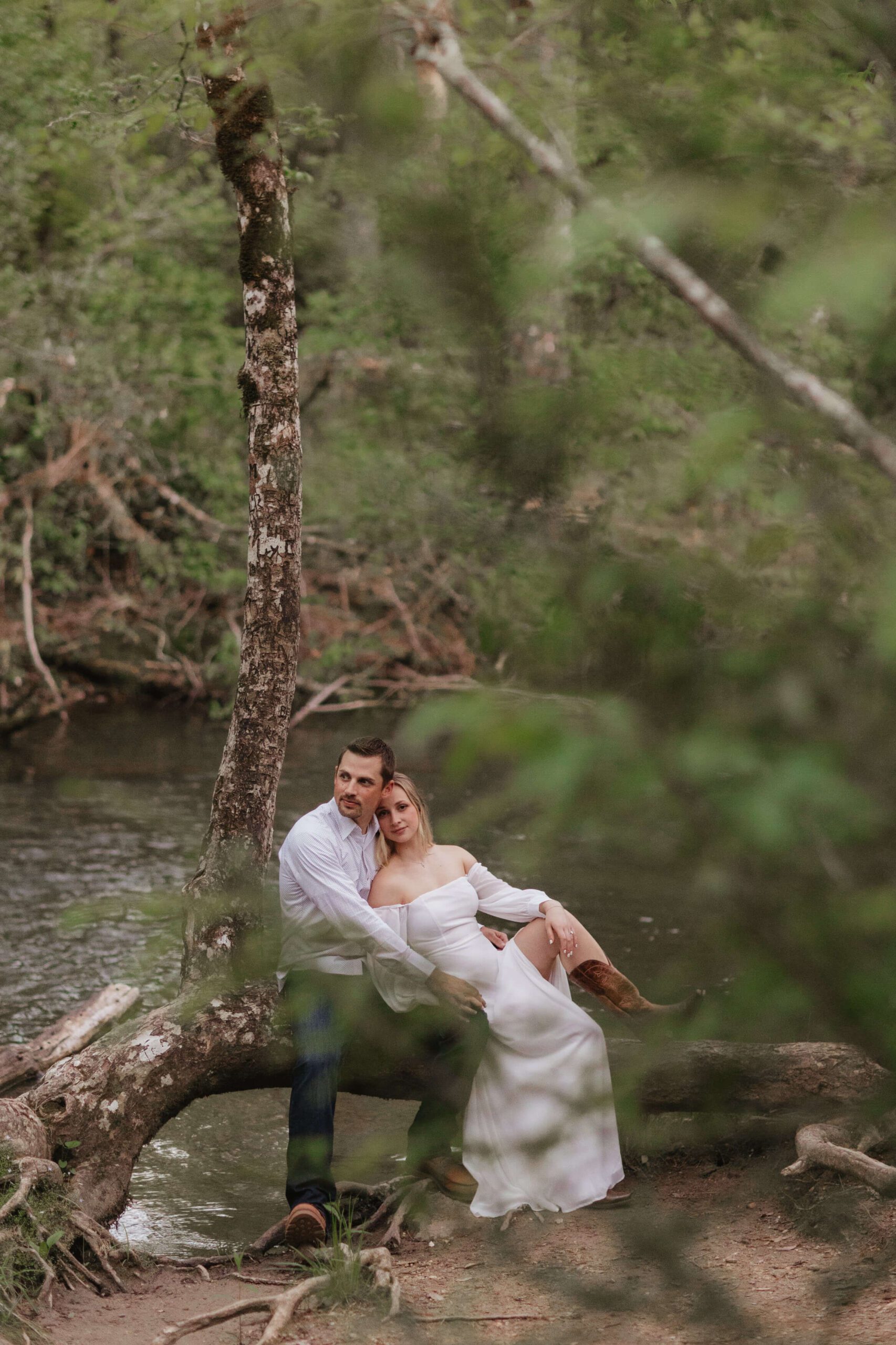 A couple sits on a fallen tree by a river in a forest. They are dressed in white and brown boots, with the woman leaning on the mans shoulder. The setting is serene, with greenery and water surrounding them.