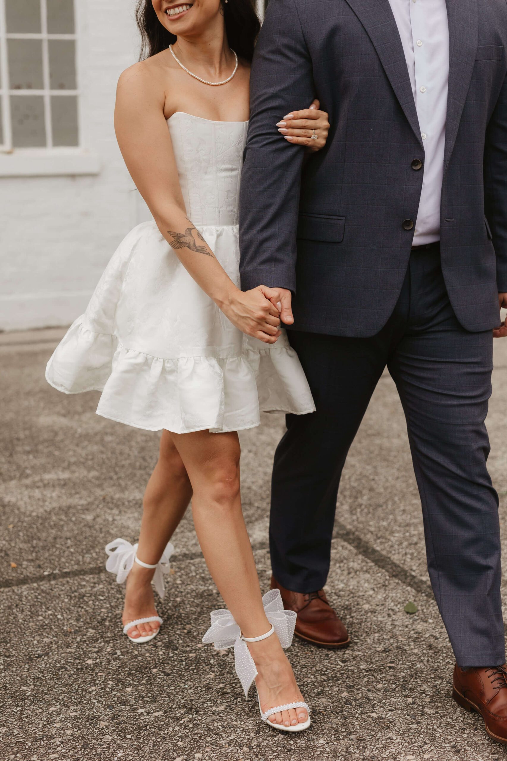 A couple walks arm in arm. The woman wears a strapless white dress with a ruffled skirt and high heels with bows. The man is dressed in a dark suit with brown shoes. They are outside on a paved walkway.