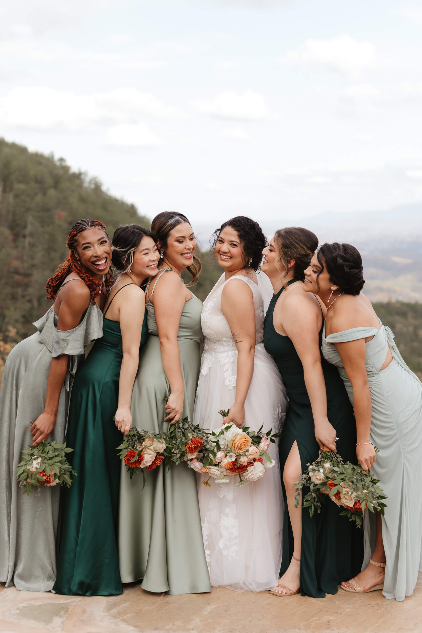 A bride in a white dress stands with five bridesmaids in green dresses, each holding bouquets of flowers. They pose smiling on a scenic overlook with a mountainous background, creating a joyful wedding moment.